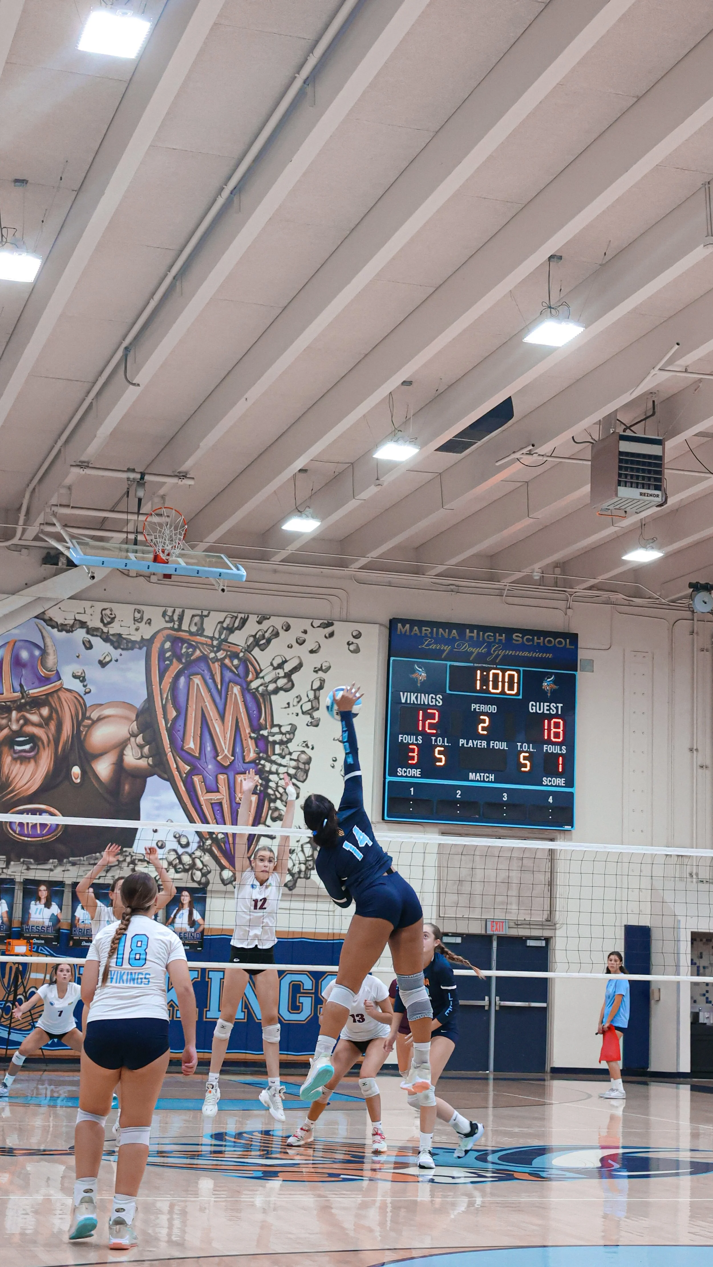 A volleyball game in progress at Marina High School with players jumping to hit the ball, a digital scoreboard showing Vikings leading 12-18, with 1 minute remaining on the clock.
