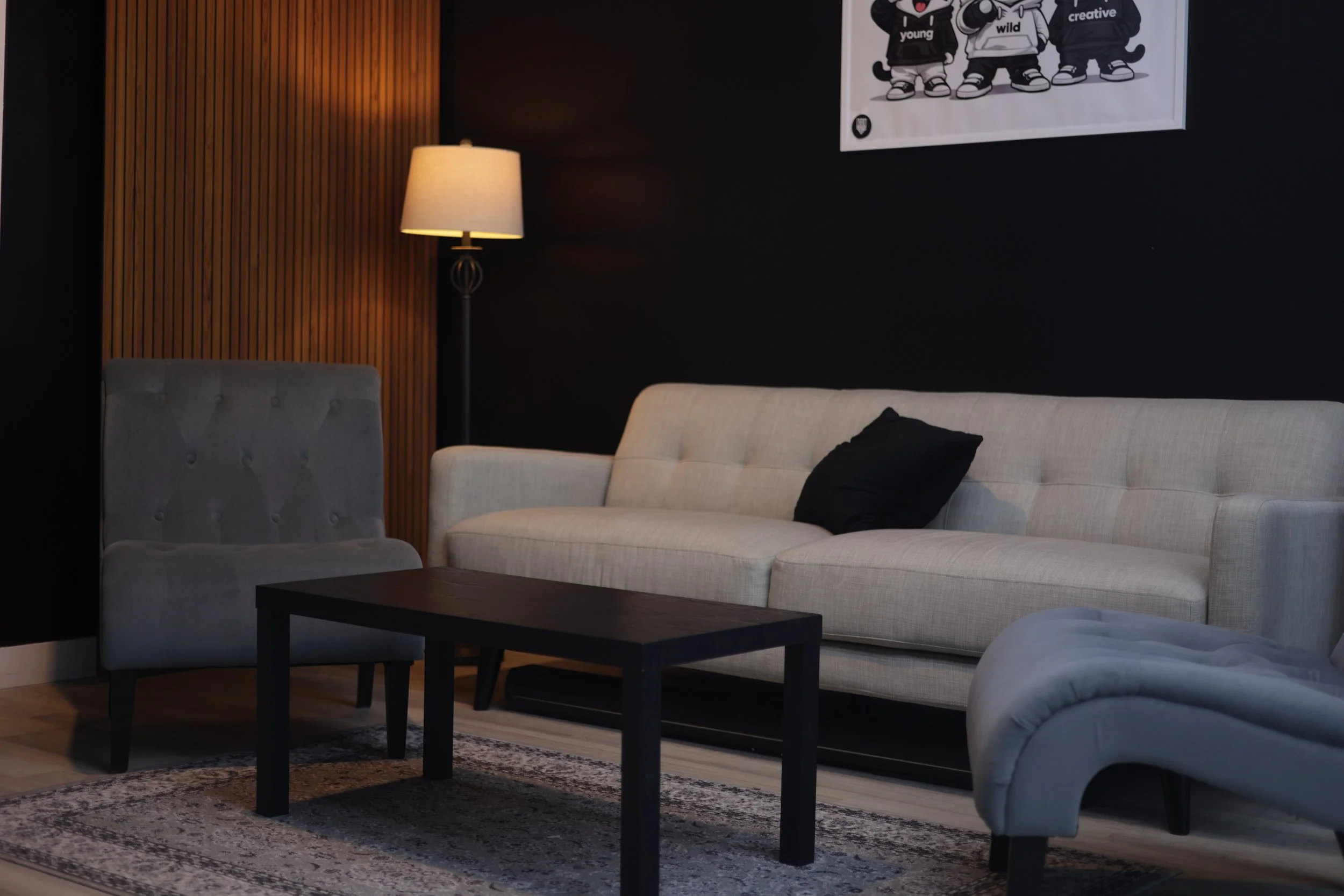 Living room with a white sofa, a grey chair, a black coffee table, a wooden panel wall, a tall floor lamp, a patterned area rug, and a poster of three cartoon characters.