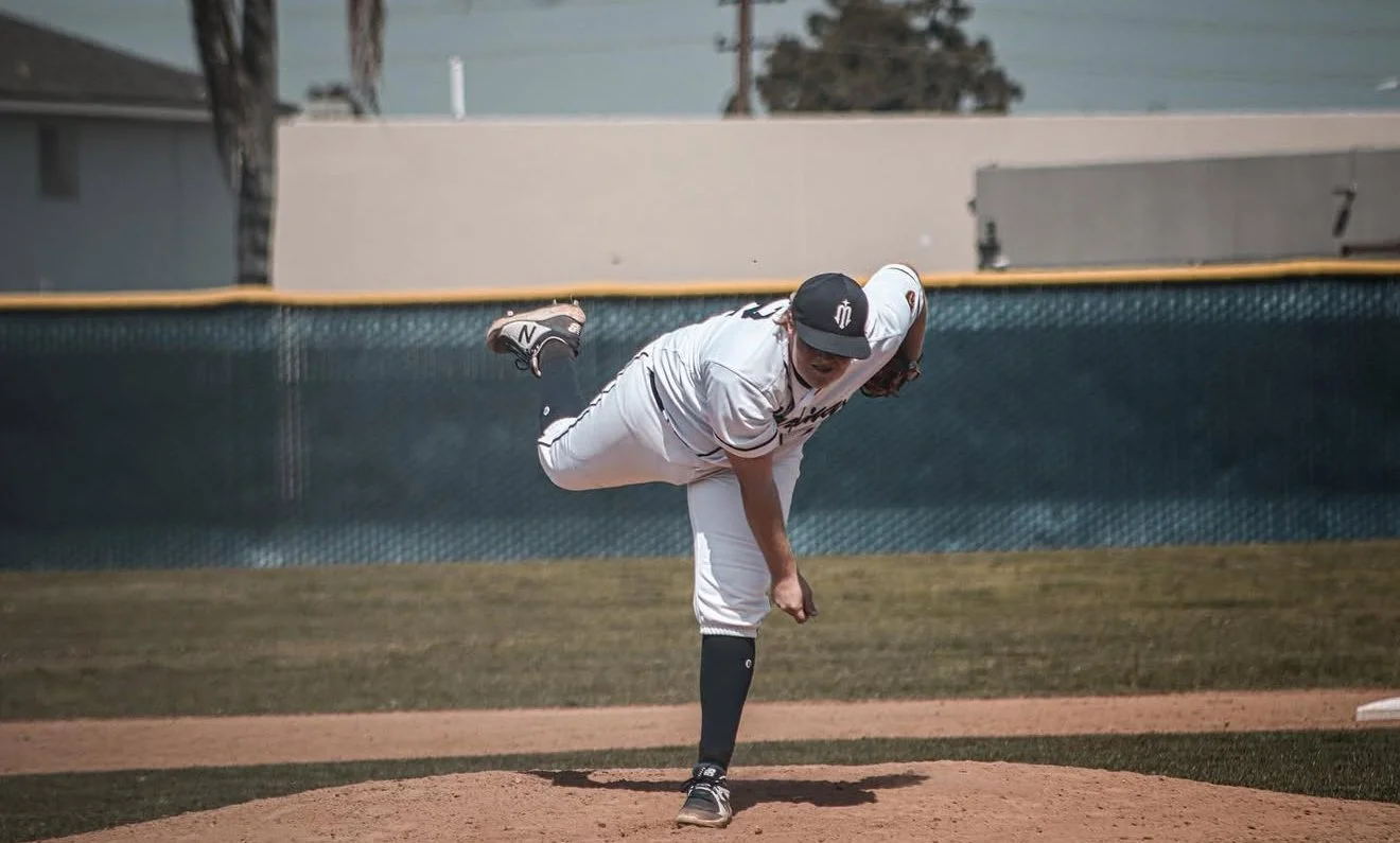 A baseball player in a white uniform and black cap pitches from the mound. The player is in a dynamic pitching stance with one leg lifted and arm extended.