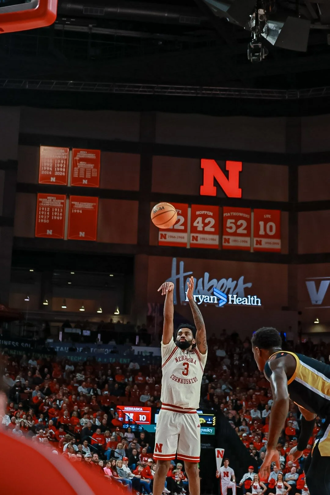 A basketball player from Nebraska Cornhuskers wearing jersey number 3 is taking a free throw in a crowded indoor arena. The background shows red banners with retired jersey numbers and a large red 'N' sign. Fans are seated in the stands, observing th