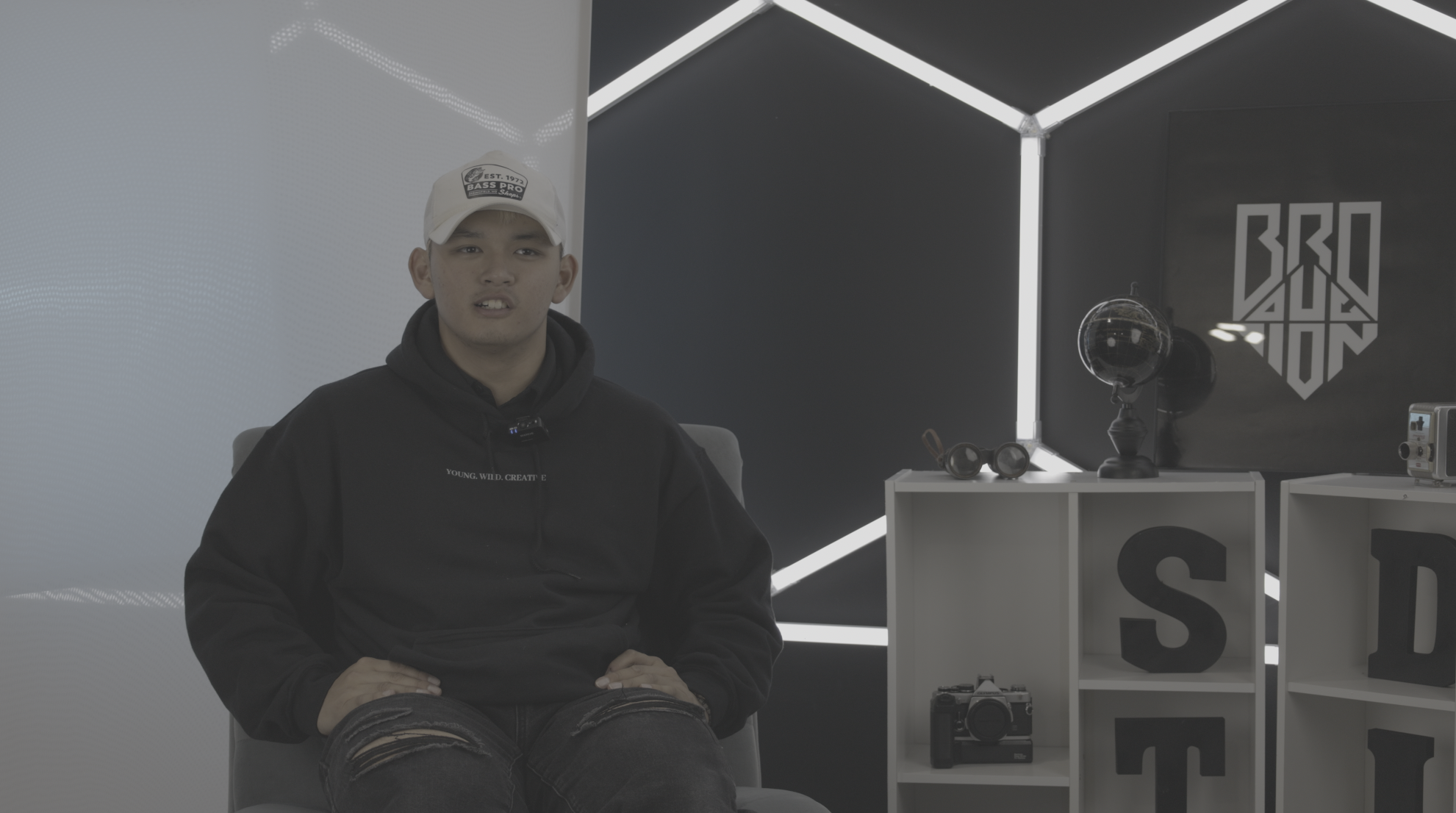 Young man sitting in a modern room with a black and white color scheme, wearing a white baseball cap and a black hoodie. Behind him are shelves with various objects, including a globe, headphones, and cameras.