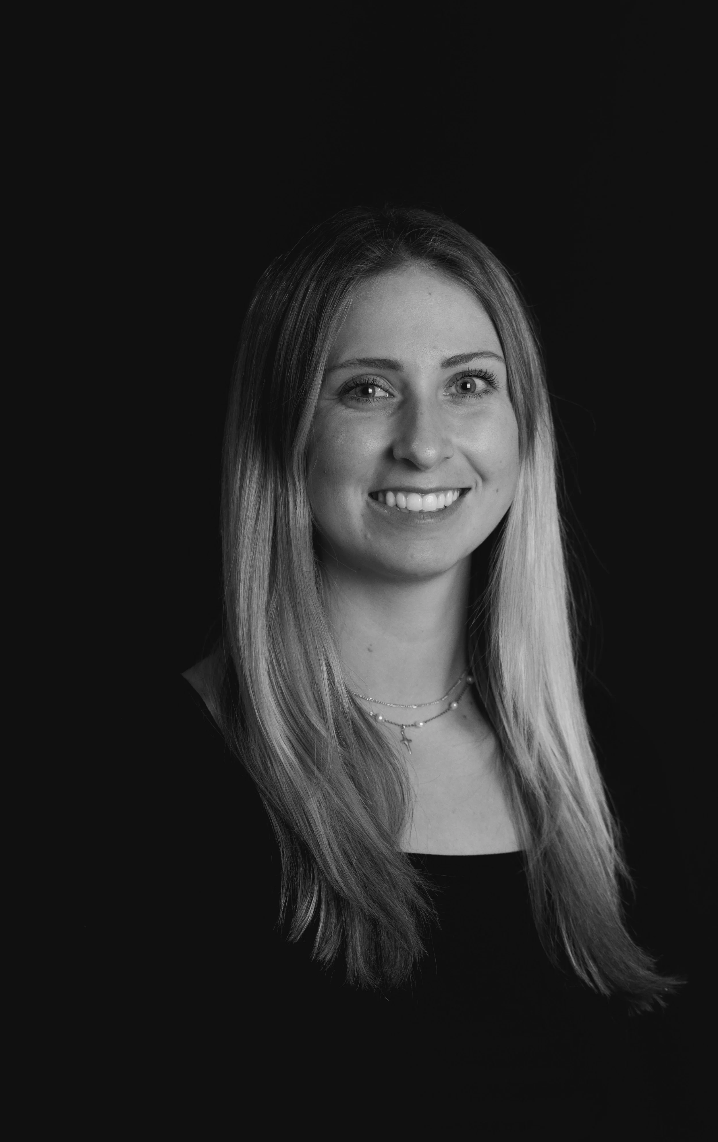 Black and white portrait of a young woman with long, straight hair, smiling, wearing a dark top and layered necklaces.