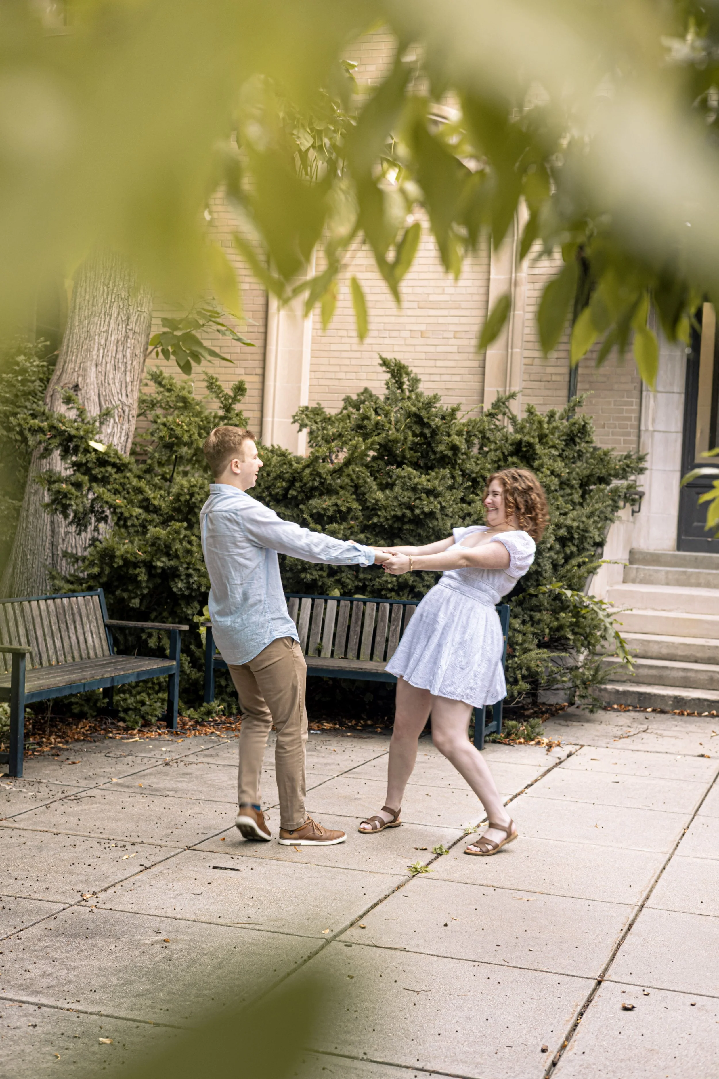 A young man and woman are holding hands and dancing on a sidewalk, surrounded by greenery and benches, in front of a building.