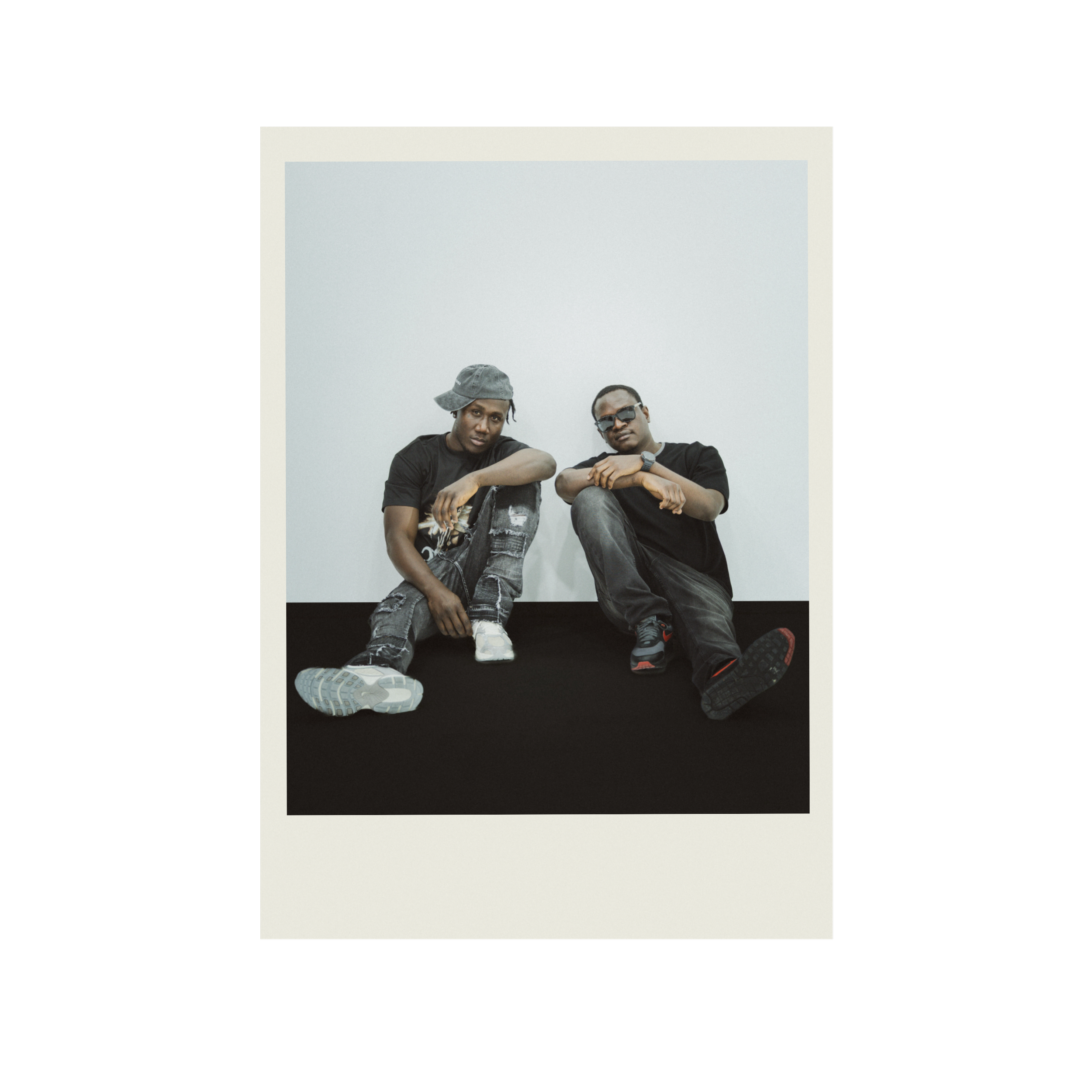 Two men sitting on the ground, one wearing a gray bucket hat, and both dressed in dark T-shirts and jeans, with one man wearing sunglasses, against a plain, light background.