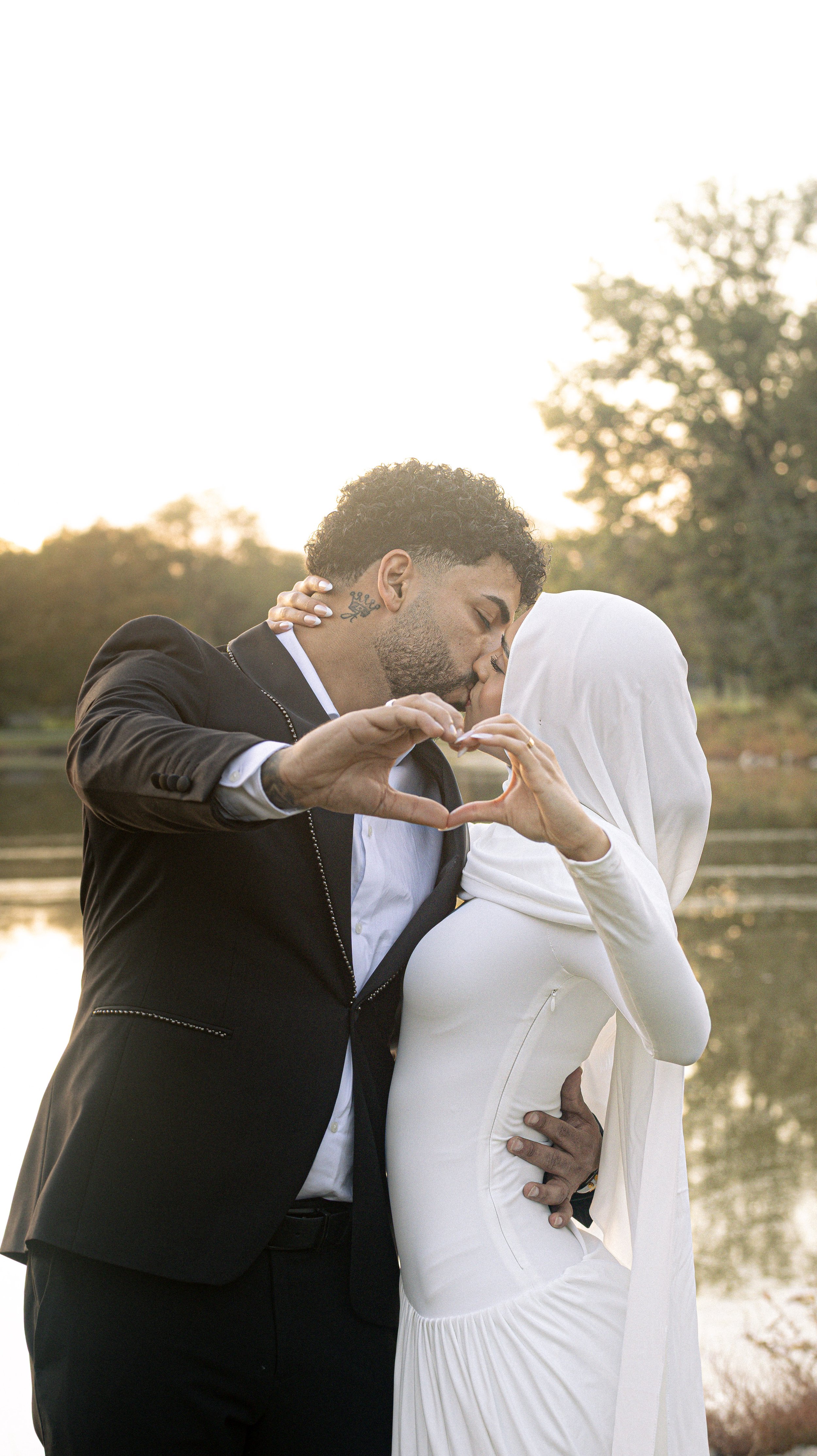 A couple sharing a kiss by a lake at sunset, making a heart shape with their hands.