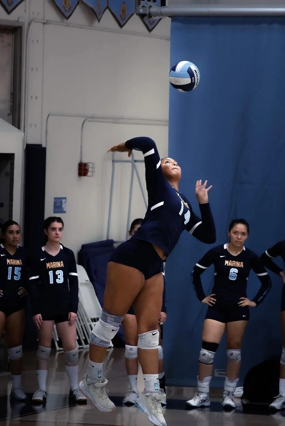 A female volleyball player in a navy blue uniform with the number 12 spikes a volleyball in an indoor gym, with her teammates watching.