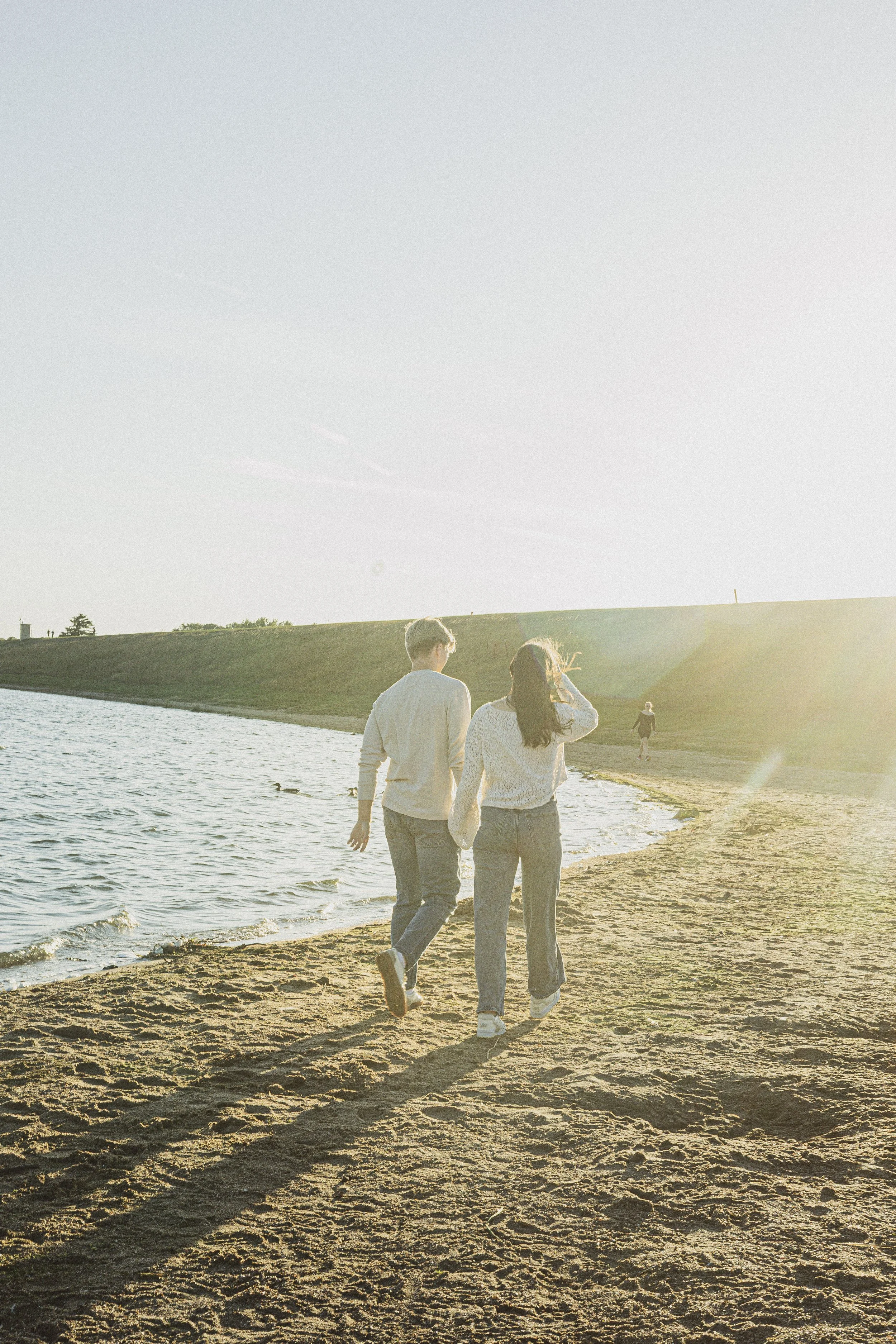 A couple walking along a sandy beach at sunset, with a grassy hill in the background and a person walking further away in the distance.