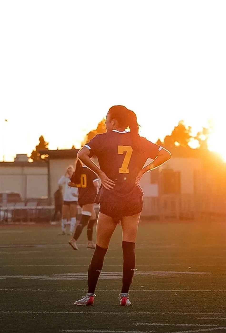 A female soccer player in a navy uniform with the number 7 stands on a soccer field at sunset, with her hands on her hips. Other players and a person are visible in the background.