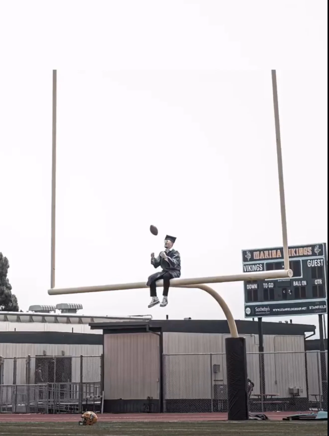 A person sitting on a football goalpost at a stadium, dressed in graduation attire, tossing a football into the air.