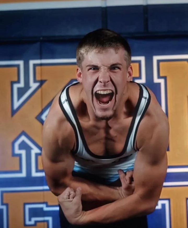Young man with short hair making an angry face, flexing his biceps, wearing a tank top, in front of a large blue and gold sports banner.