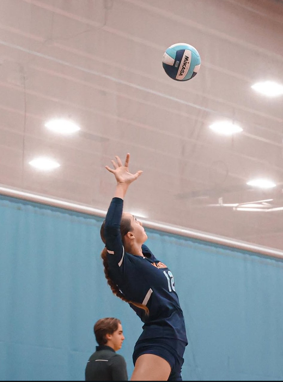 A female volleyball player wearing a dark uniform jumps to hit a volleyball inside an indoor gym with a blue wall and a ceiling with multiple lights.