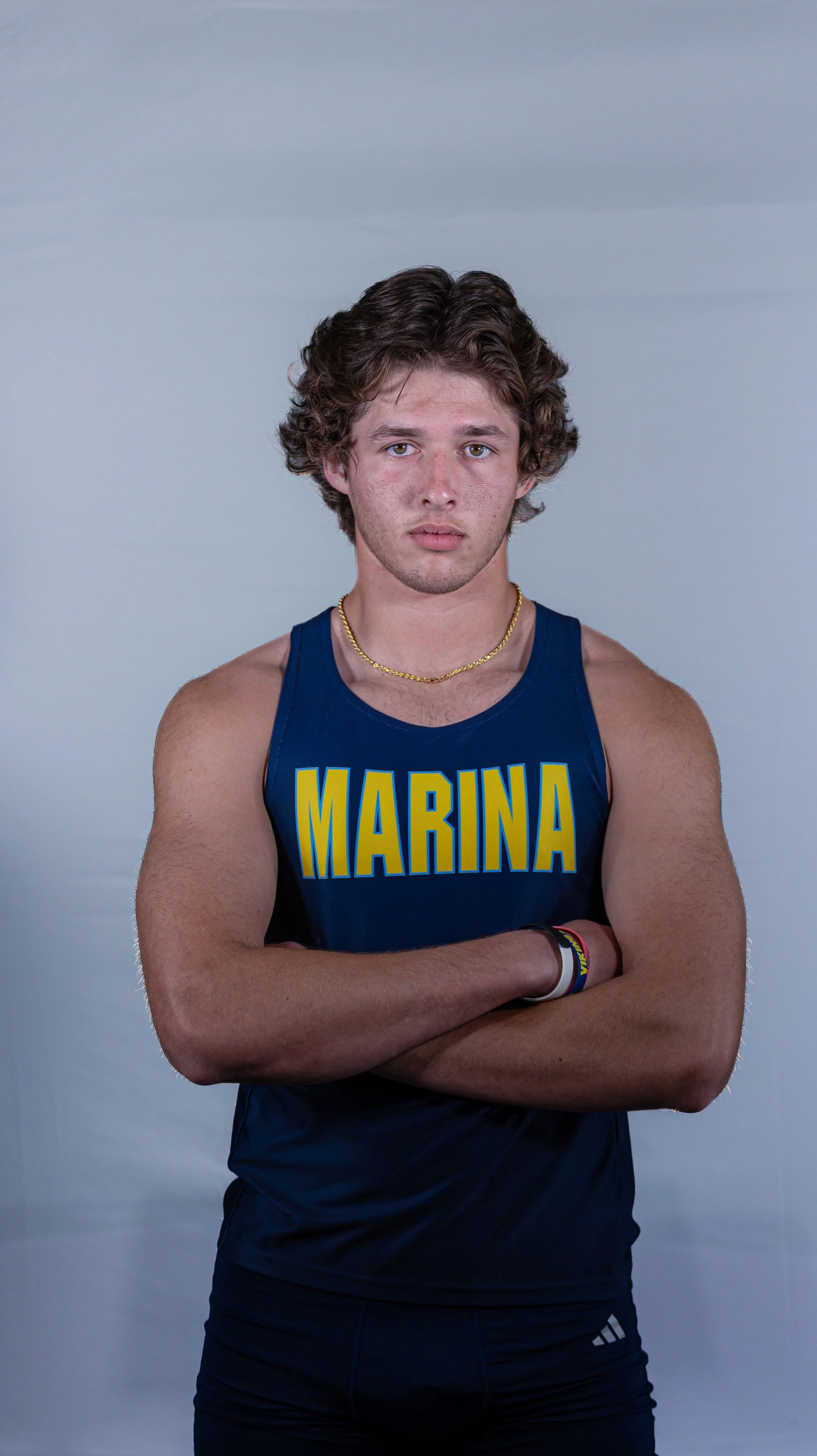 A young male athlete with curly brown hair, standing with arms crossed, wearing a blue tank top with the word "MARINA" in yellow letters and black athletic shorts.