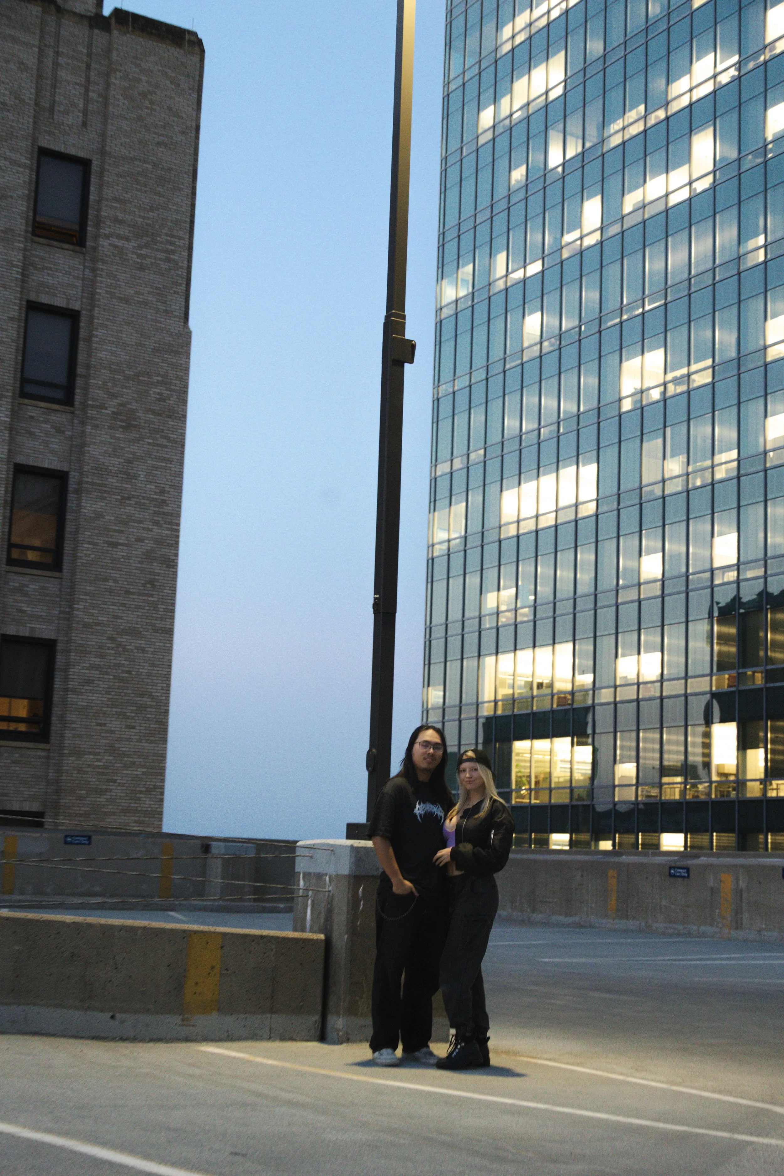 A man and woman standing together in an urban parking garage, with tall buildings and reflective glass windows in the background, during the evening.