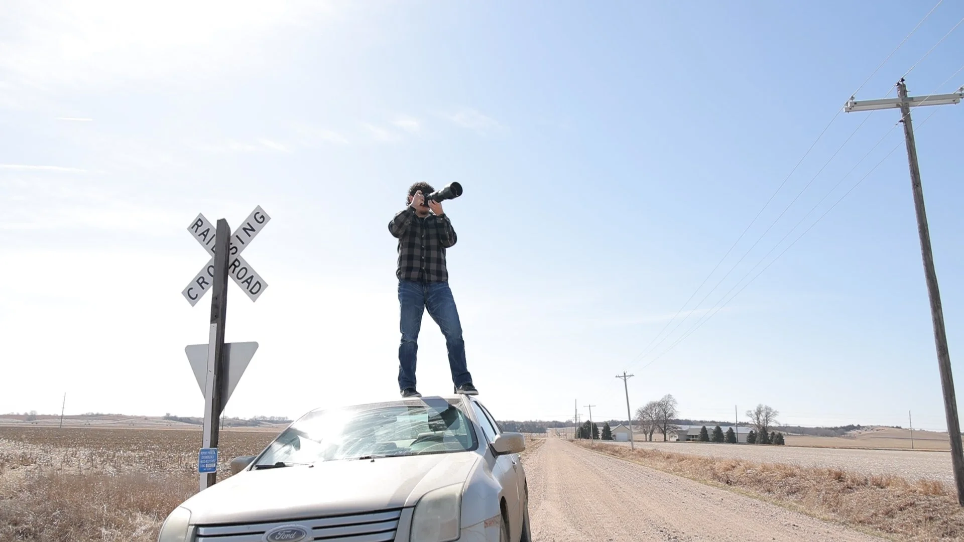 A man standing on top of a car with a camera, taking a photo in a rural area with fields and power lines, a railroad crossing sign nearby, under a clear blue sky.
