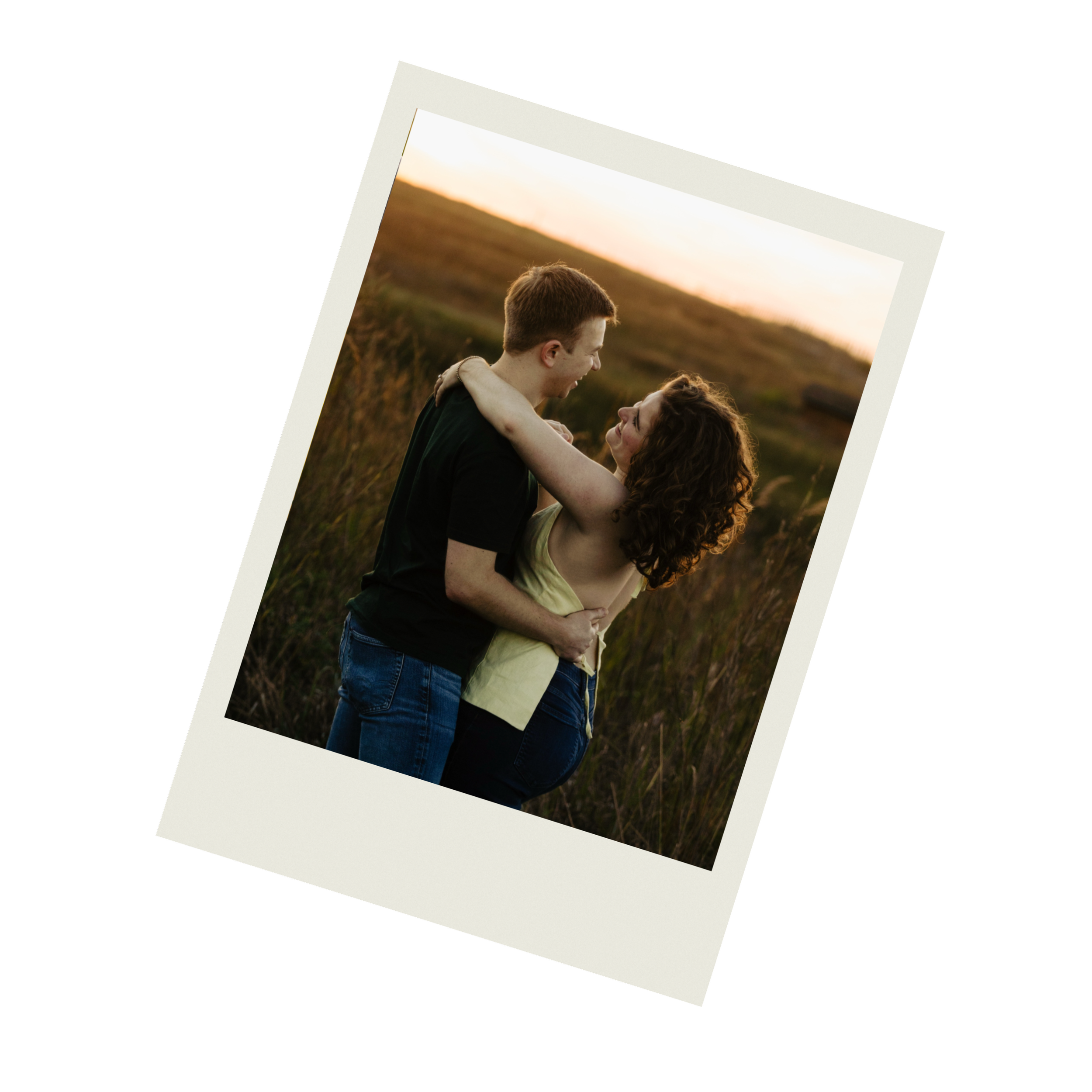 A young man and woman smiling and cuddling in a field during sunset.