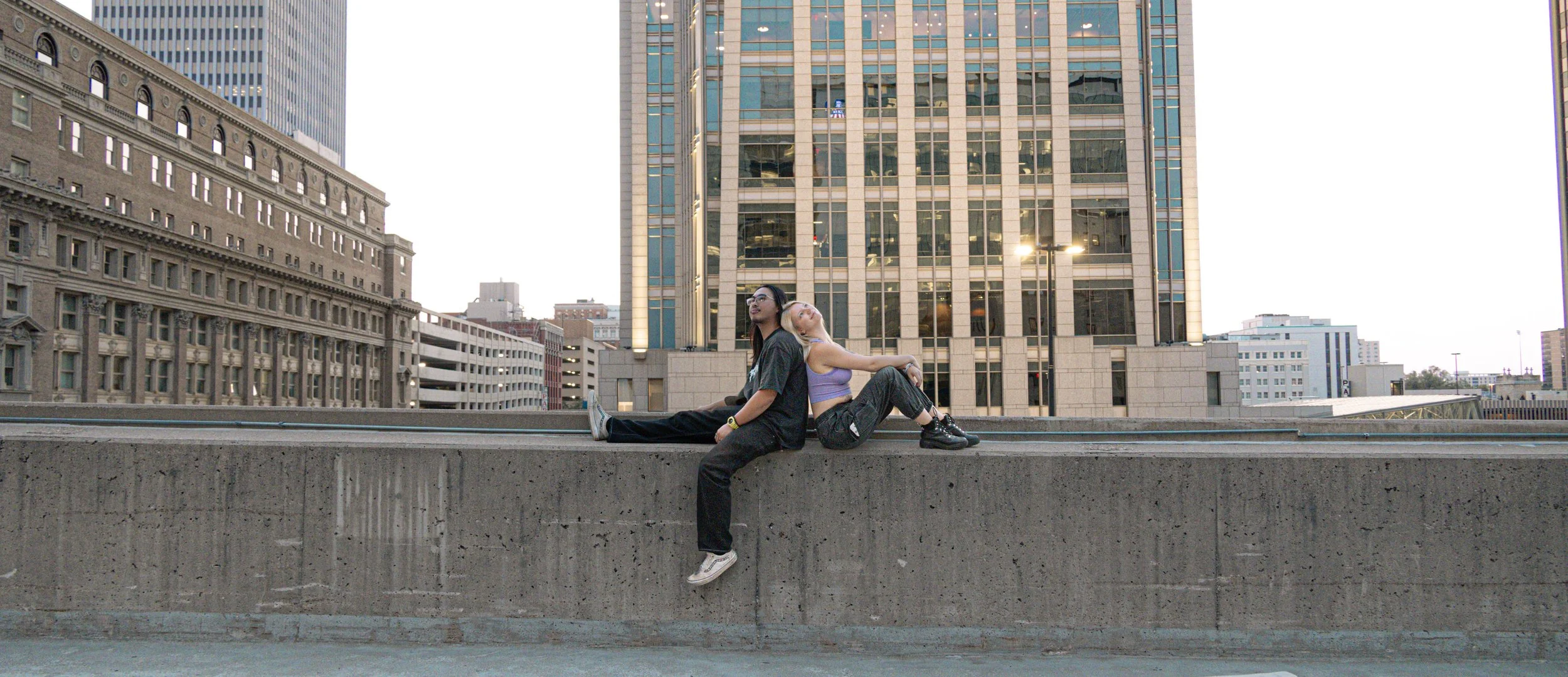 Two people sitting back-to-back on a concrete ledge in an urban setting with tall buildings in the background. The person on the left is wearing glasses, a dark shirt, and dark pants, while the person on the right is wearing a light purple top and da