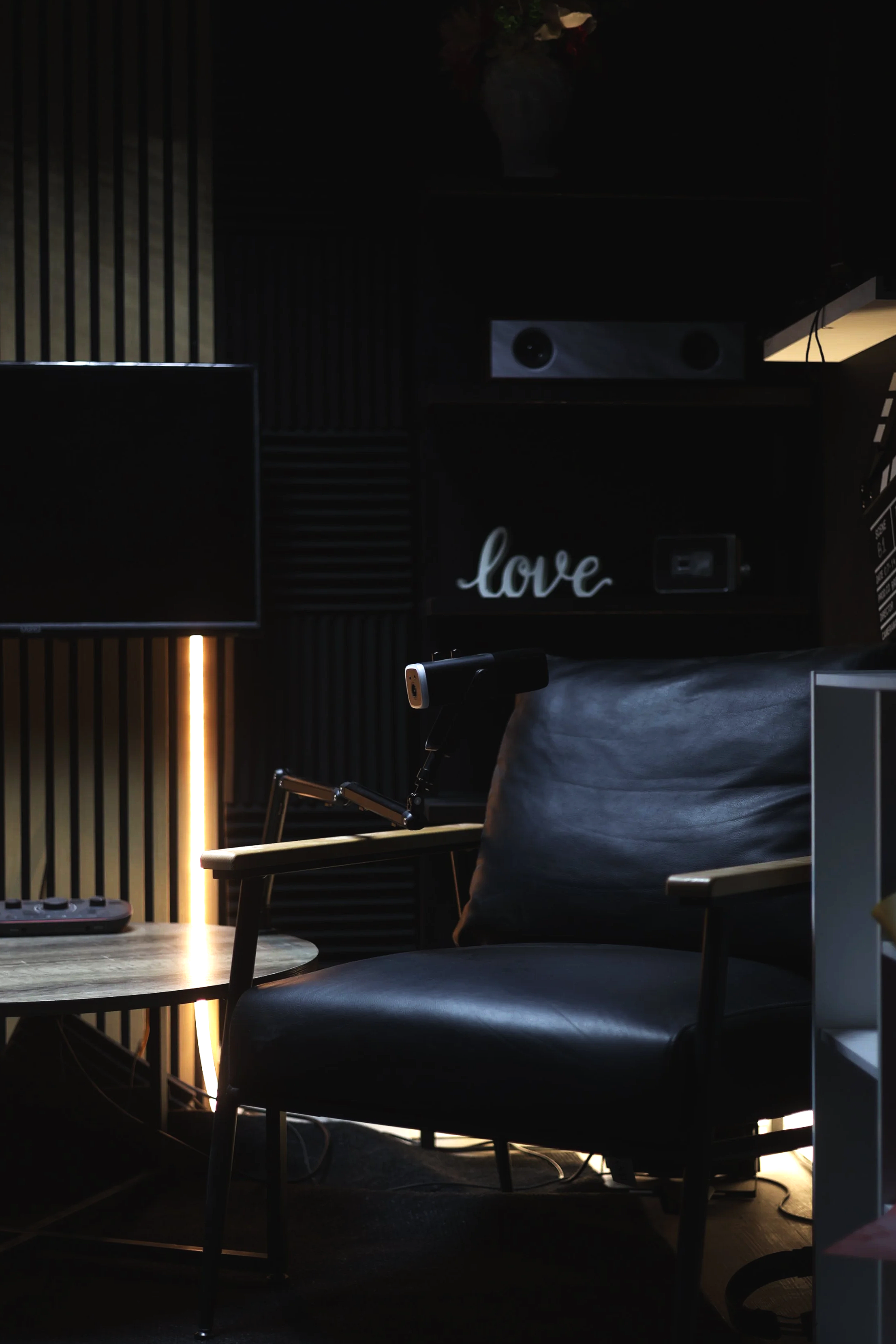 A dimly lit room with a black and wooden patterned wall, a black desk with a piece of electronic equipment, a chair with wooden armrests, and a black pillow with 'love' written on it. There is a television or monitor on the left and some decorative items and lighting accents.