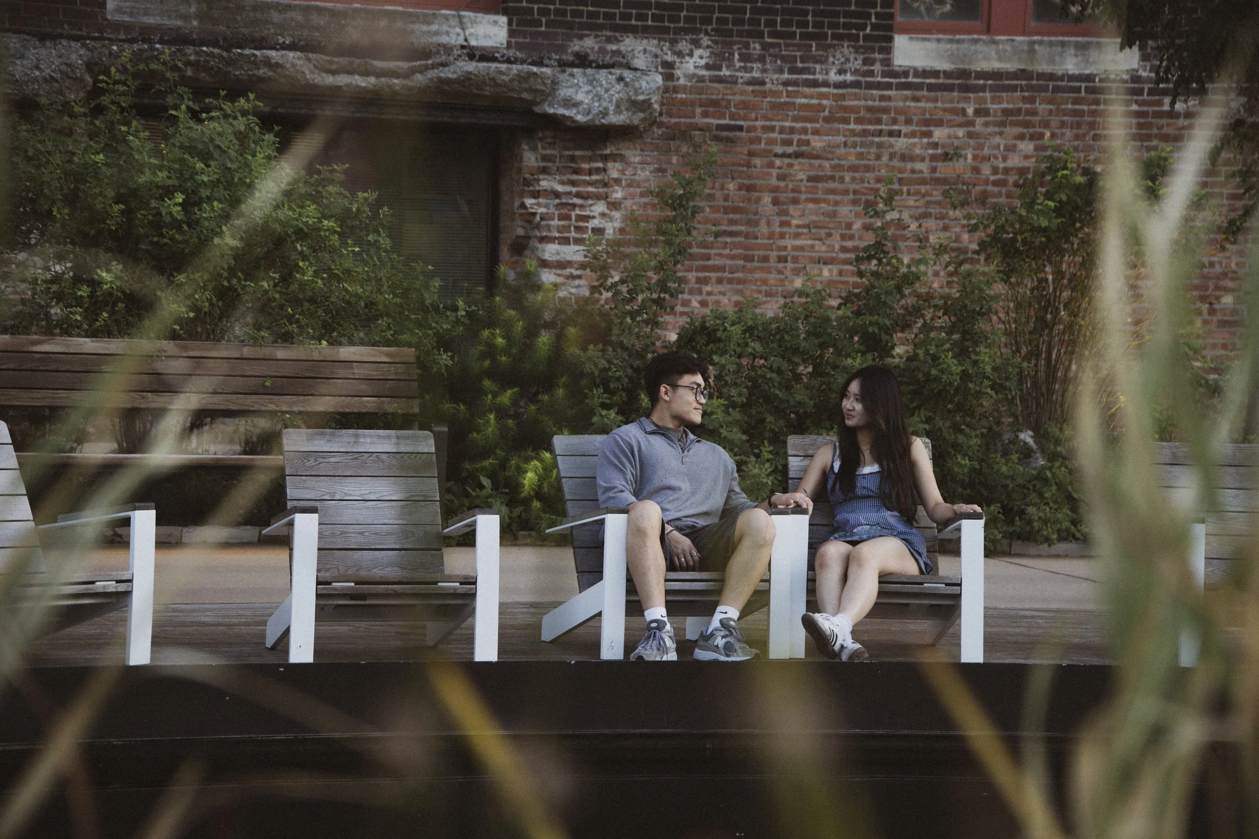 A young man and woman sitting on a bench outdoors, holding hands and looking at each other, with greenery and a brick wall in the background.