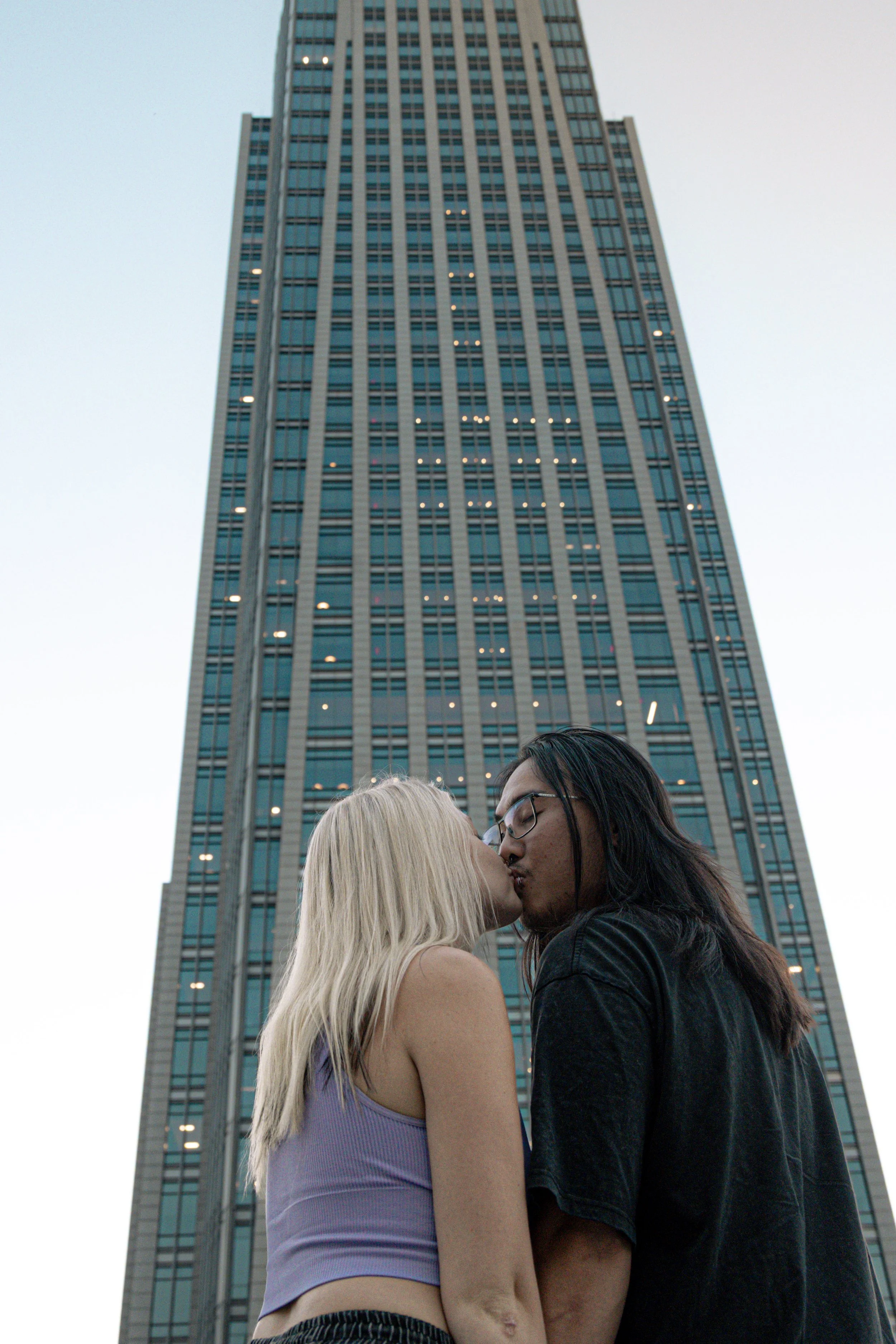 A couple kissing in front of a tall skyscraper with many lit windows at dusk.