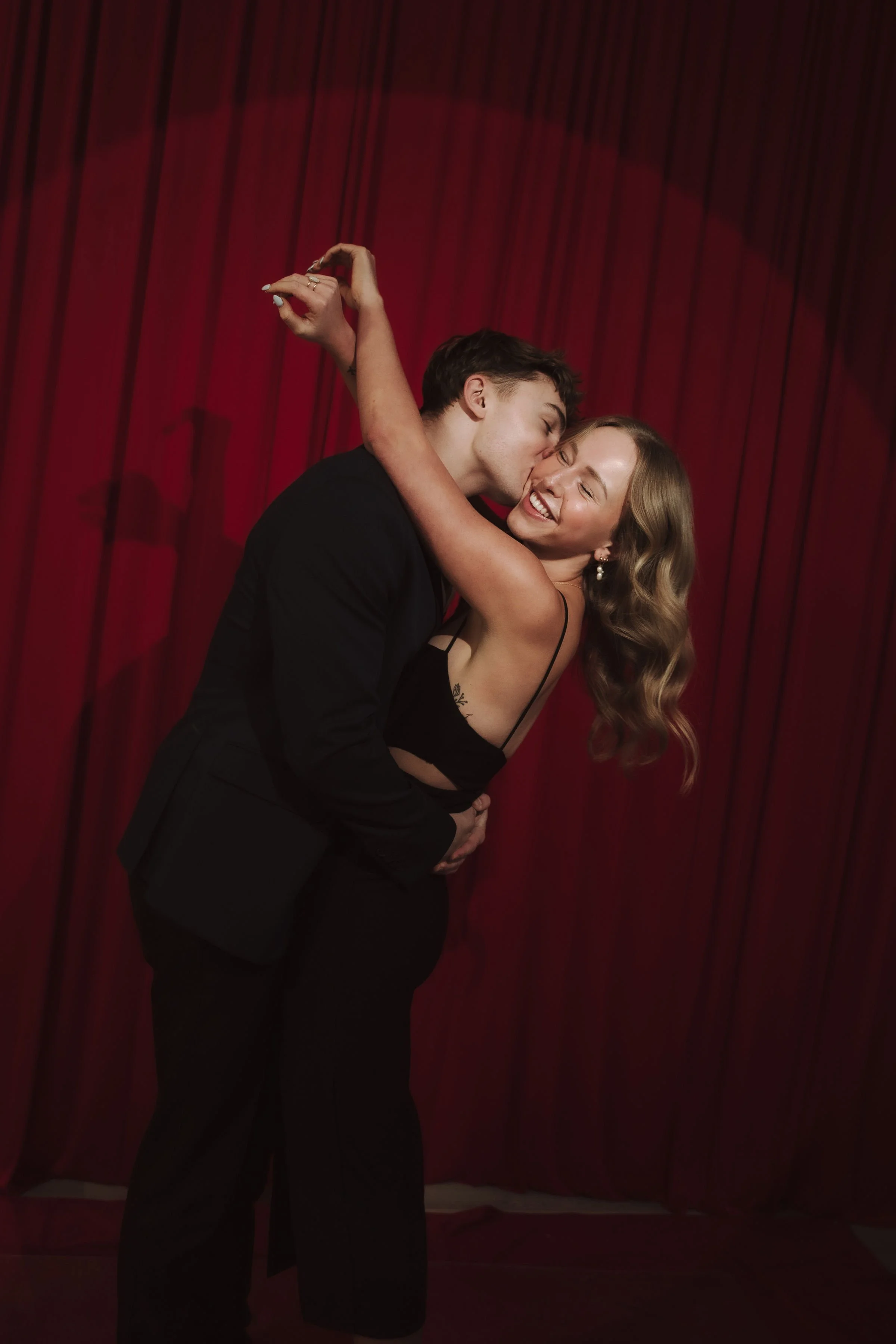 A young man and woman sharing a kiss in front of a red curtain. The man is wearing a black suit, and the woman is wearing a black dress. The woman has wavy blonde hair, and both are smiling.