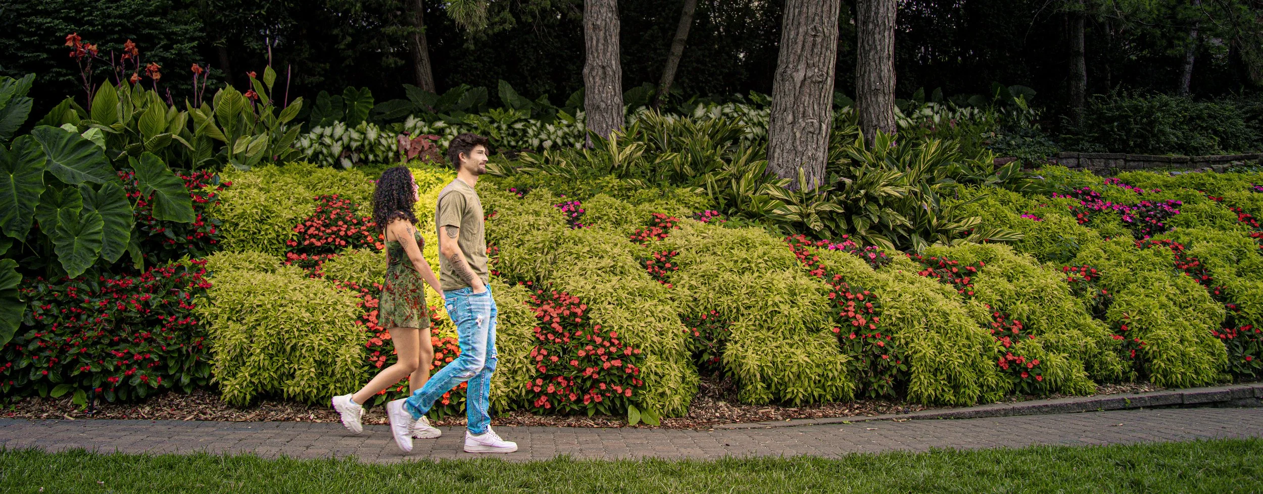 A young couple walking hand in hand on a paved path through a lush garden with green bushes, pink, red, and white flowers, and tall trees in the background.