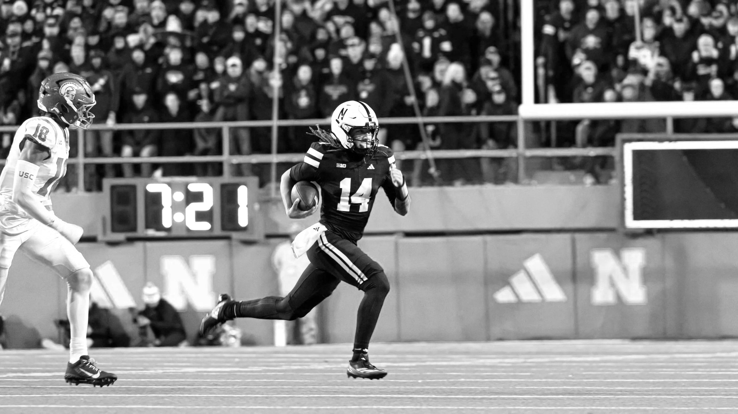 A football player wearing a dark jersey with the number 14 running with the ball on the field while an opposing player watches from behind. The background shows spectators in the stands and a scoreboard with 7:21.