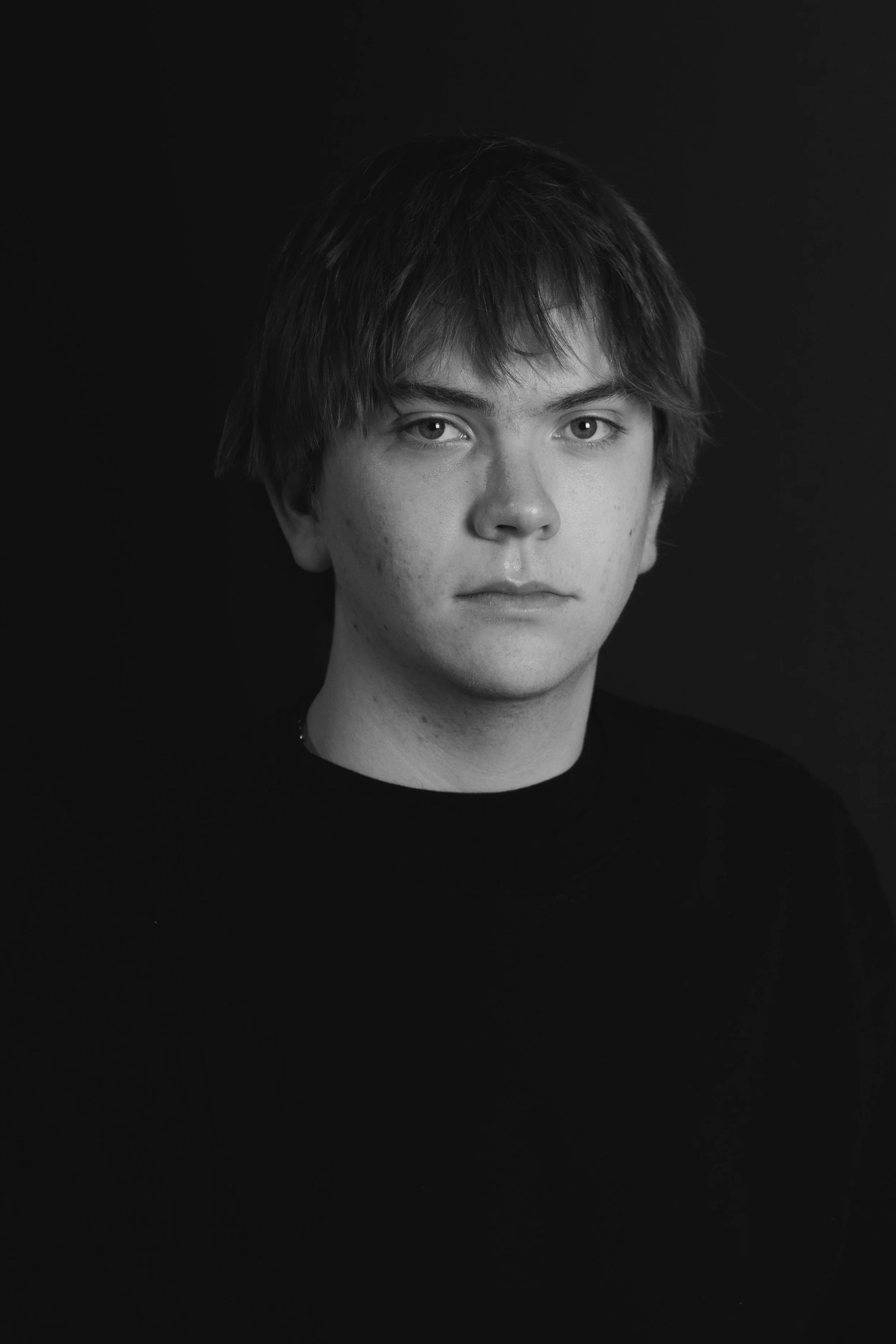 Black and white portrait of a young man with medium-length hair, looking directly at the camera against a dark background.