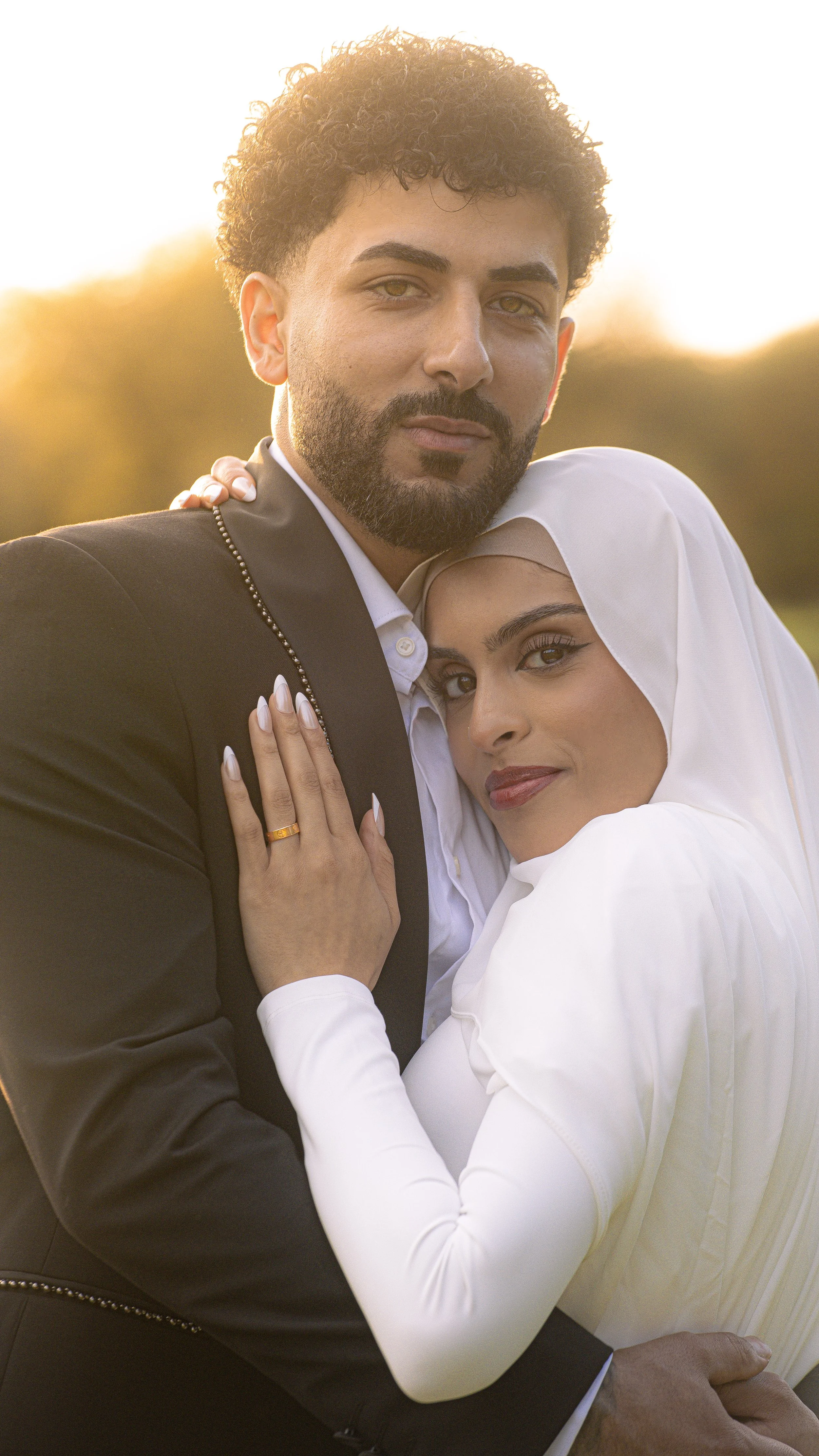 A man and woman embrace outdoors during sunset, with the woman wearing a white hijab and the man in a black suit and white shirt.