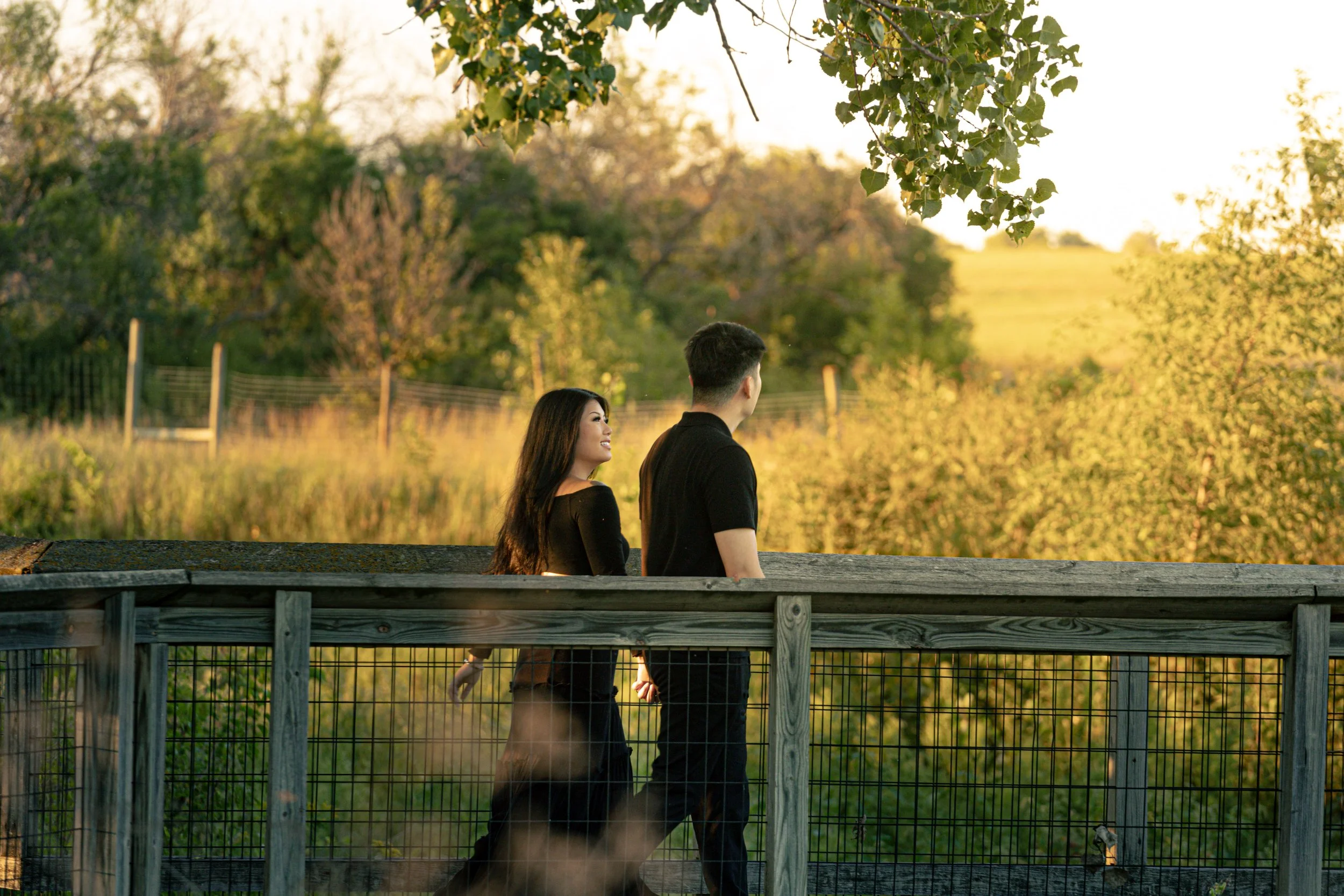 A couple holding hands walking on a wooden bridge at sunset, surrounded by trees and lush greenery.