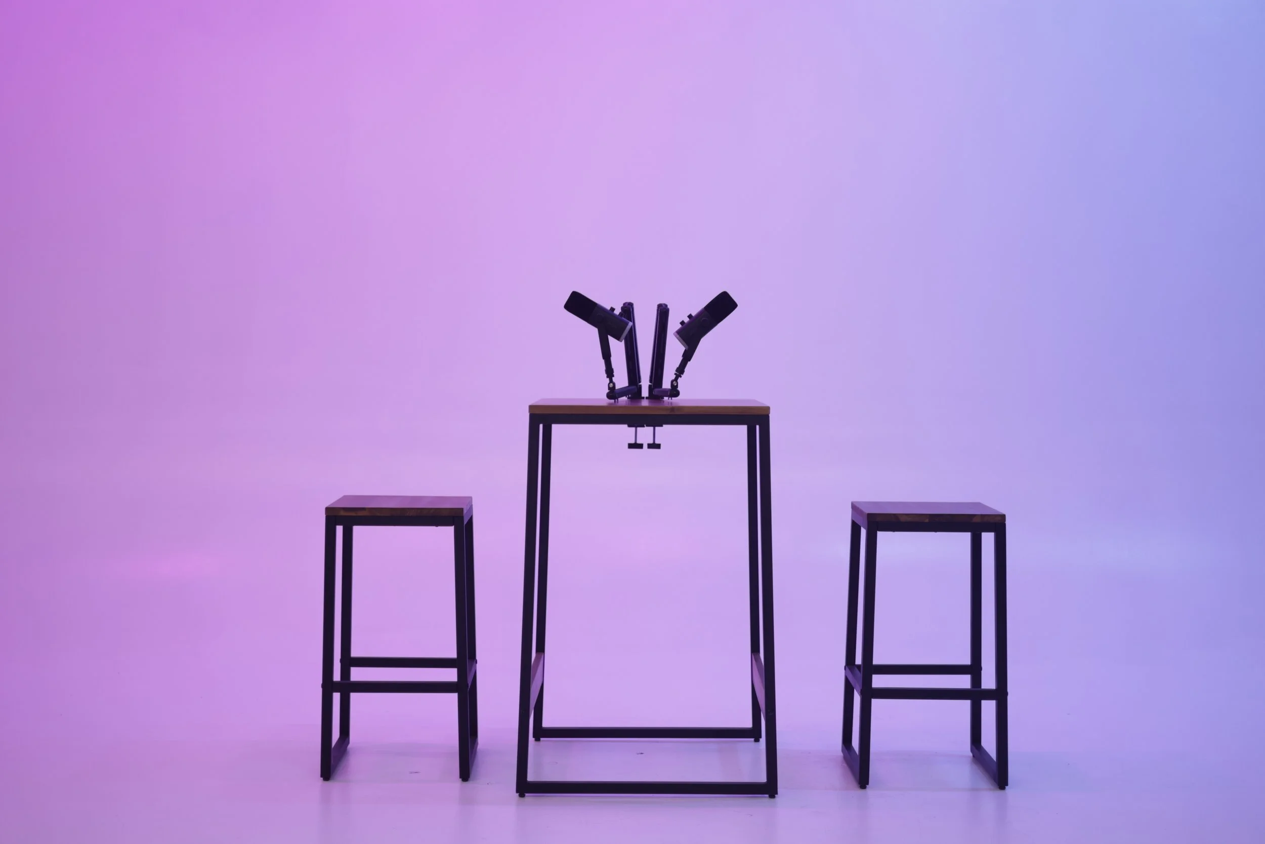 A minimalist setup with three wooden stools and a small table, set against a purple gradient background, with a mechanical device or robotic arms mounted on the table.