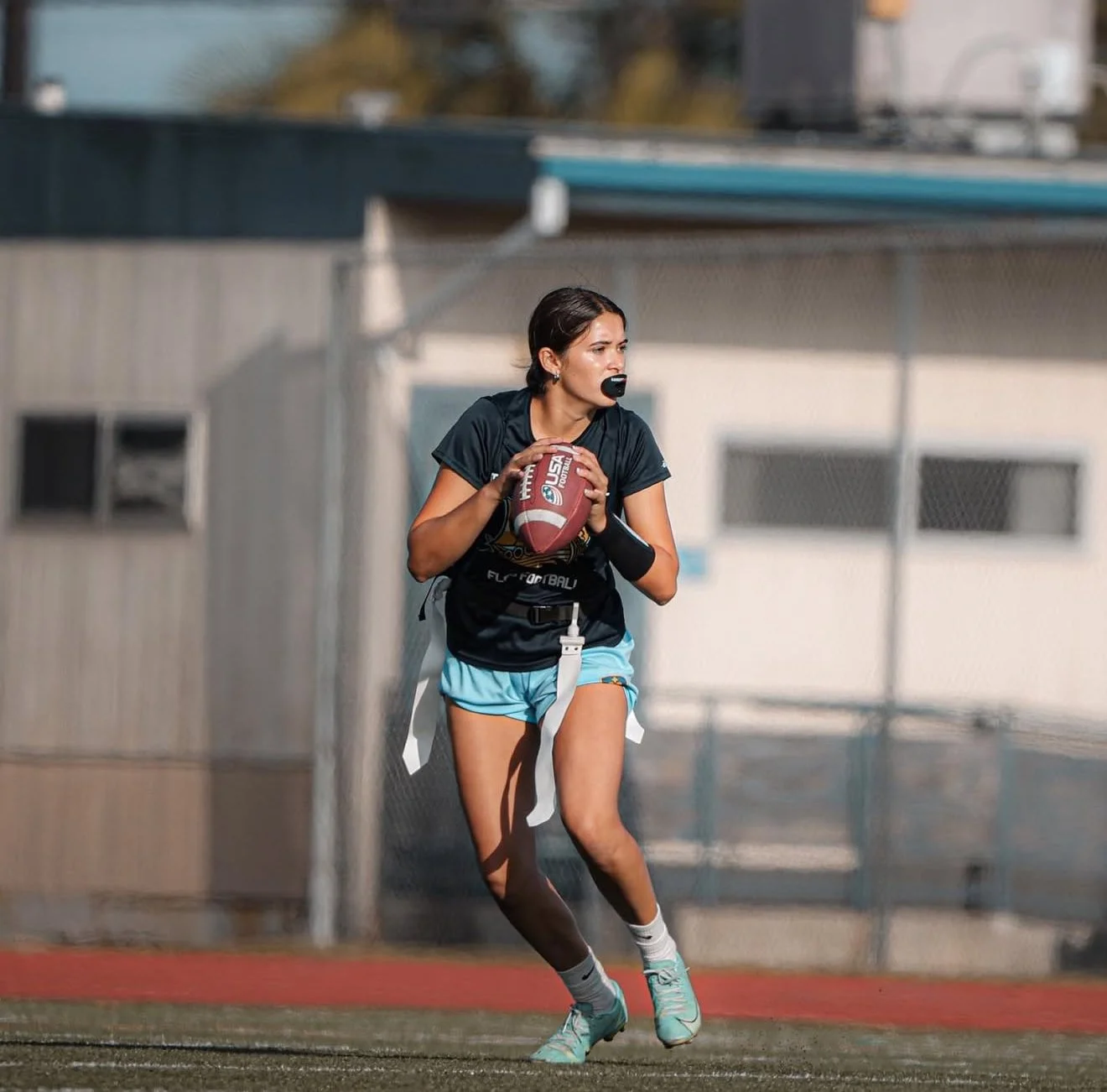 A female rugby player is holding a football on a field, wearing a black jersey, light blue shorts, and teal cleats, with a wristband on her left arm.