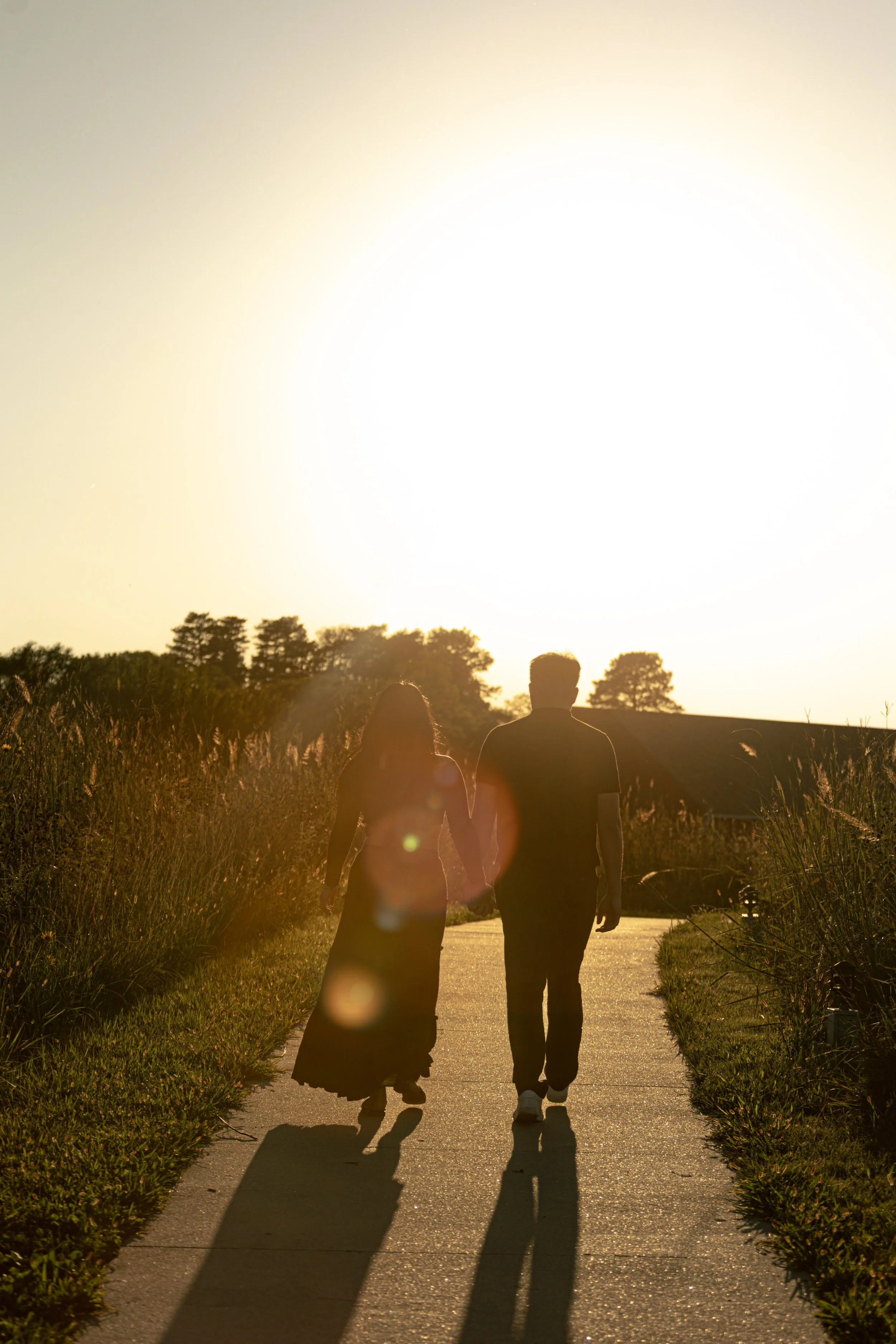 A couple walking hand in hand on a sidewalk during sunset, with tall grass on either side and a building in the background.