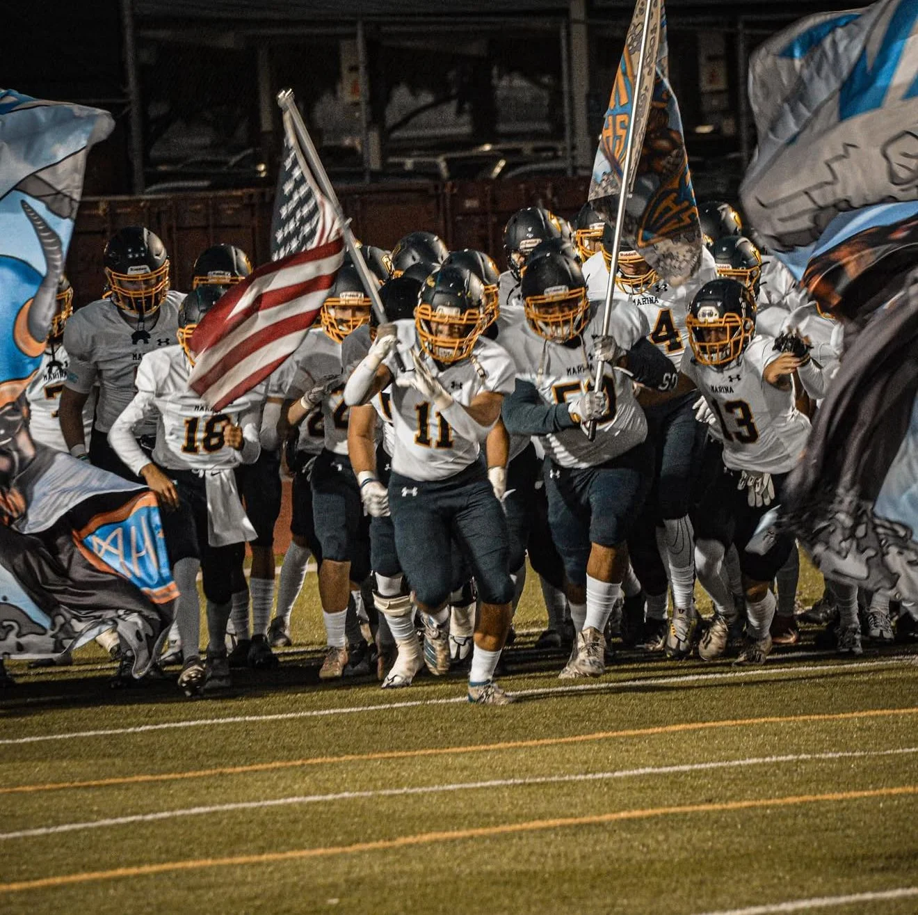High school football players running onto the field under night sky, holding flags, wearing uniforms, helmets, and cleats.