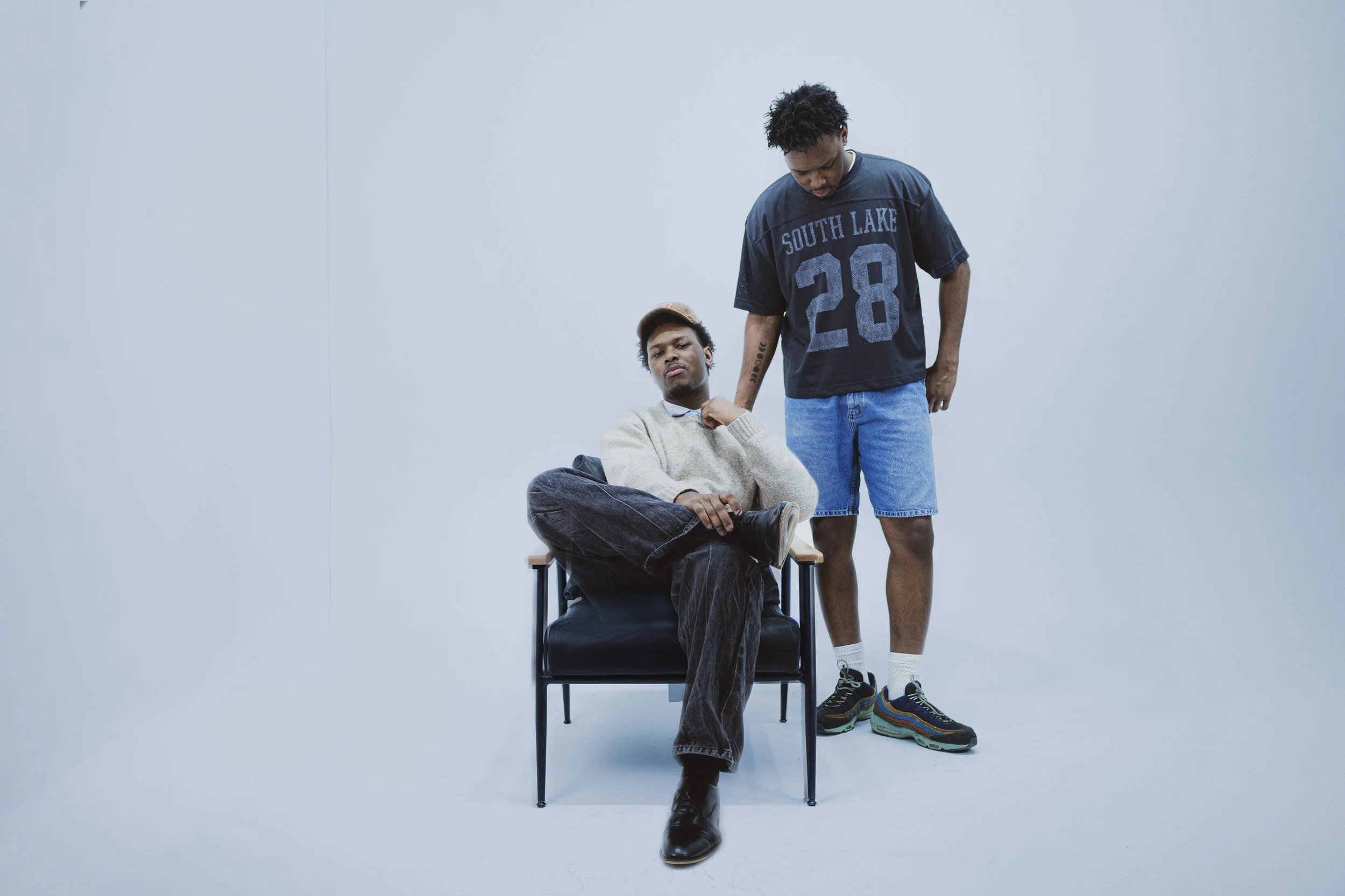 Two young men in casual clothes are in a plain white room. One is sitting on a chair with crossed legs, wearing a beige sweater, dark pants, and a beige cap. The other stands beside him, wearing a black T-shirt with 'SOUTH LAKE 28' printed on it, and