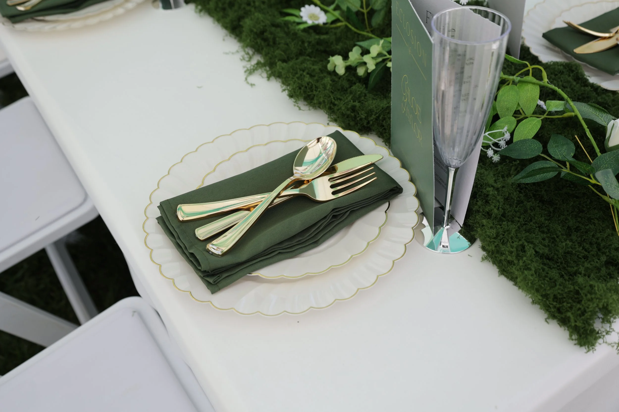 A table set for a formal event with a white tablecloth, gold and green cutlery on a folded green napkin, a white plate with a gold rim, greenery, flowers, and a tall glass of water.