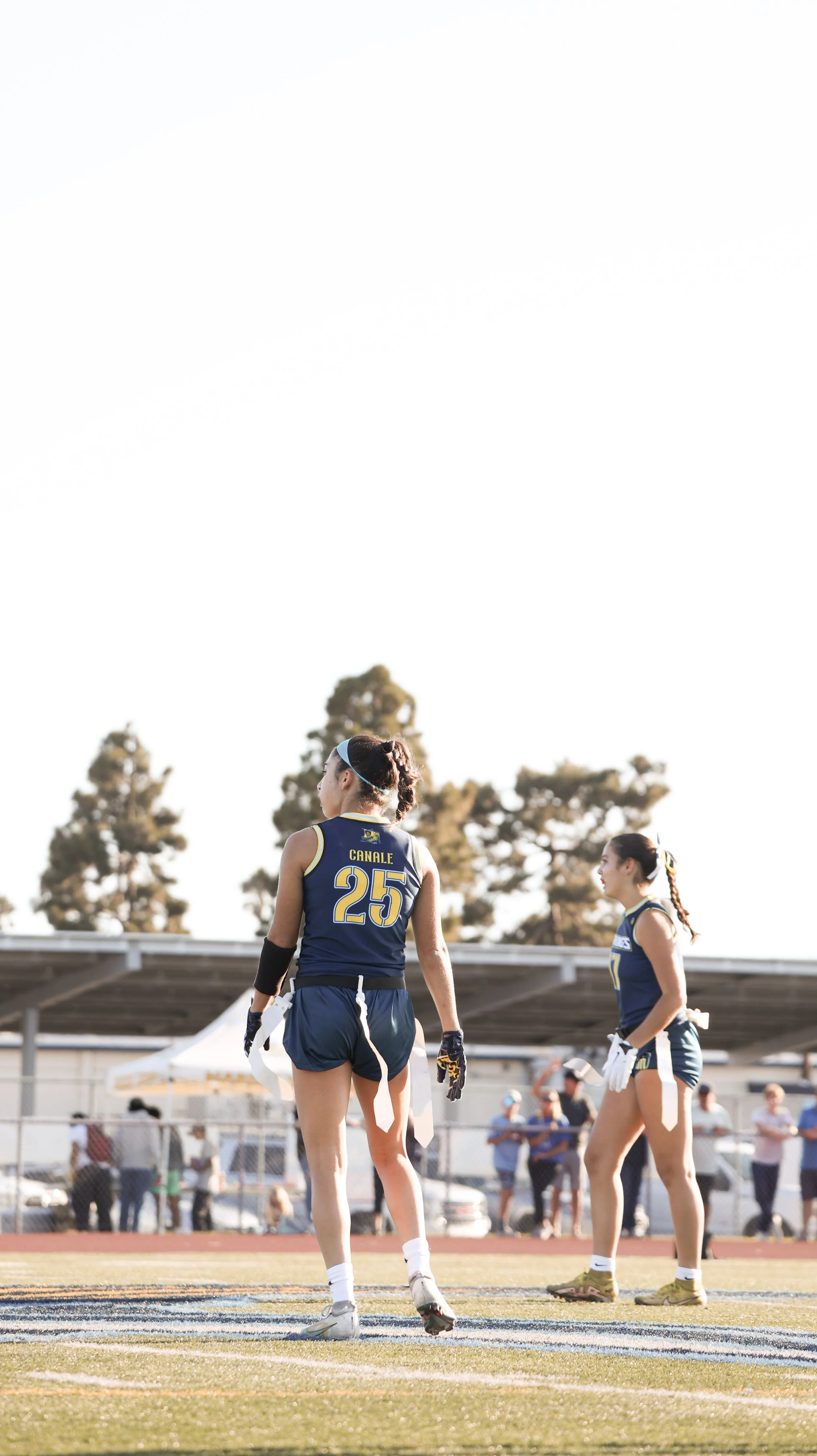 Two female track athletes in navy blue uniforms with yellow accents standing on a track during a competition, with spectators and trees in the background.