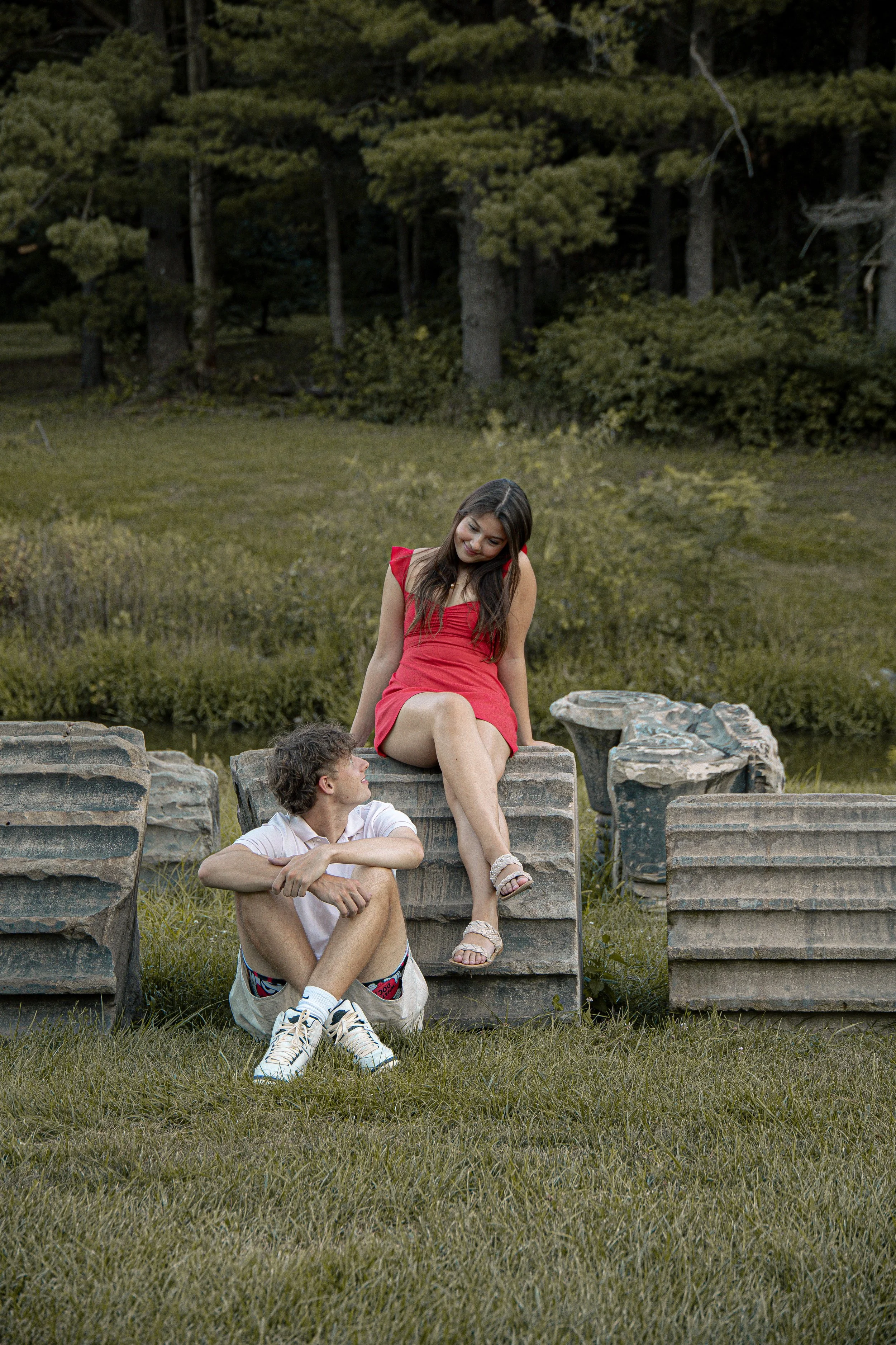 A young man sitting cross-legged on the grass looking up at a young woman sitting on a stone structure in an outdoor setting with trees in the background.