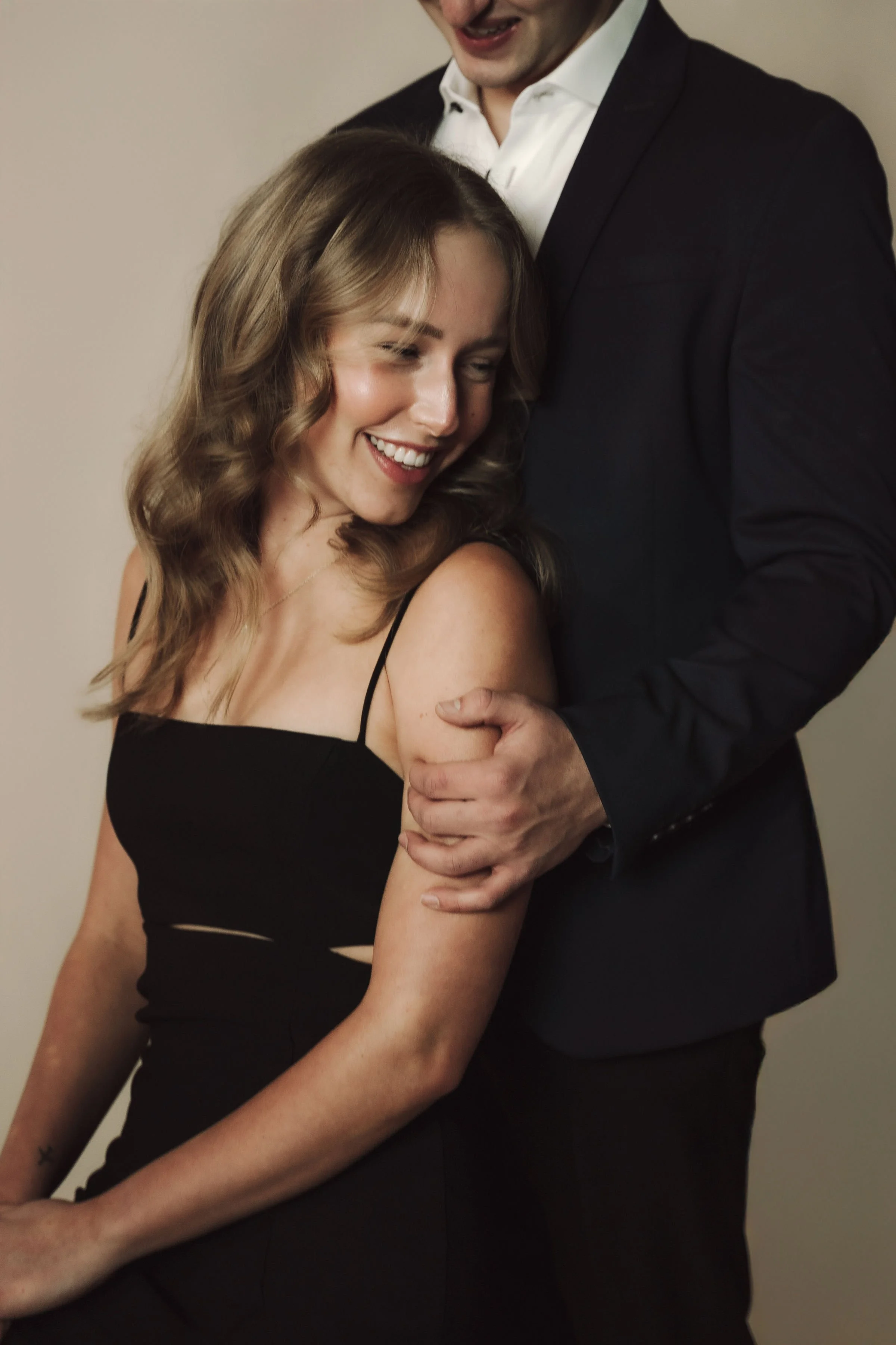 A woman with wavy brunette hair smiling and wearing a black dress, standing close to a man in a black suit and white shirt, both smiling and displaying affection.