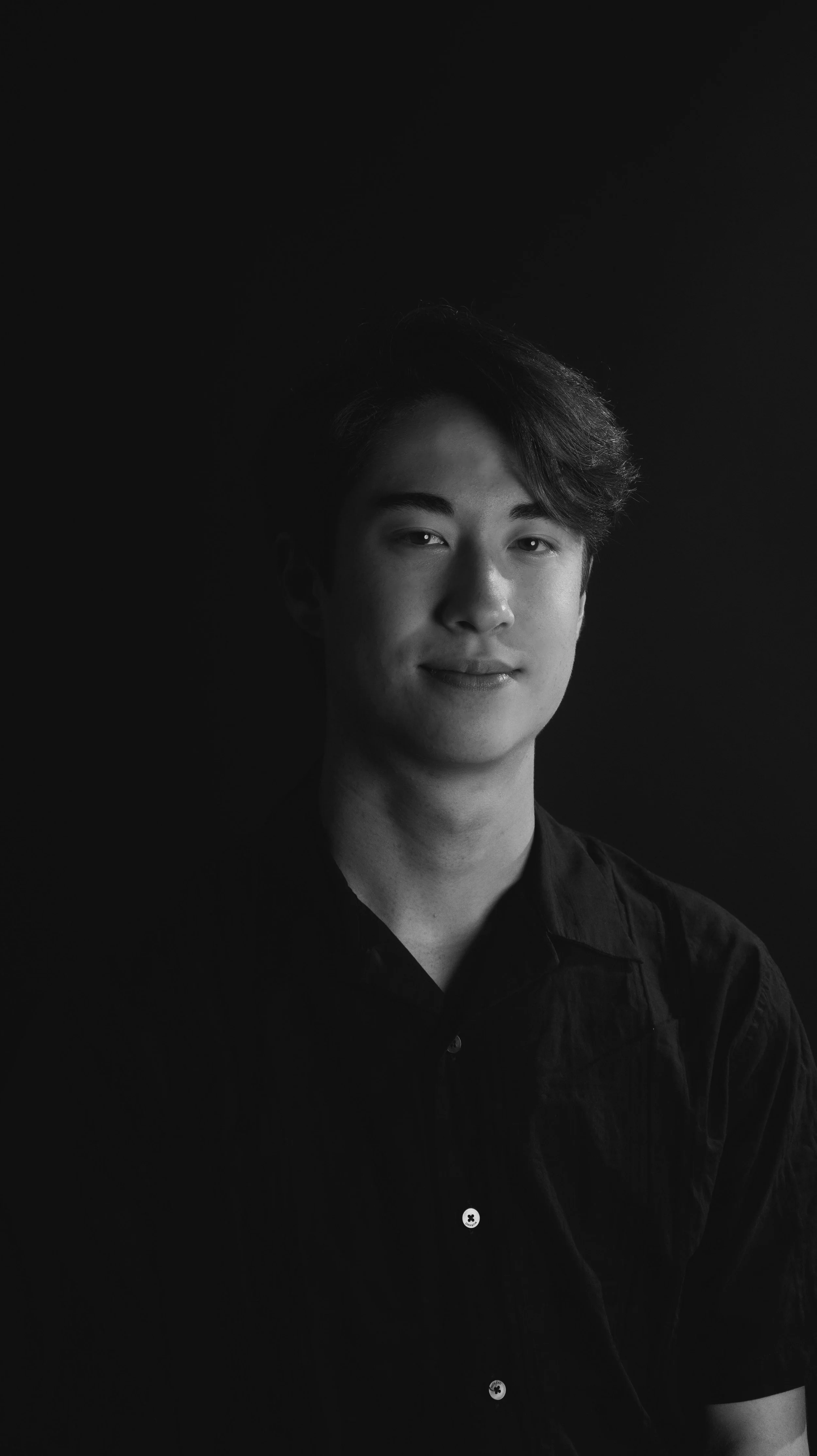 Black and white portrait of a young man with short hair, wearing a black button-up shirt, smiling slightly, with a dark background.