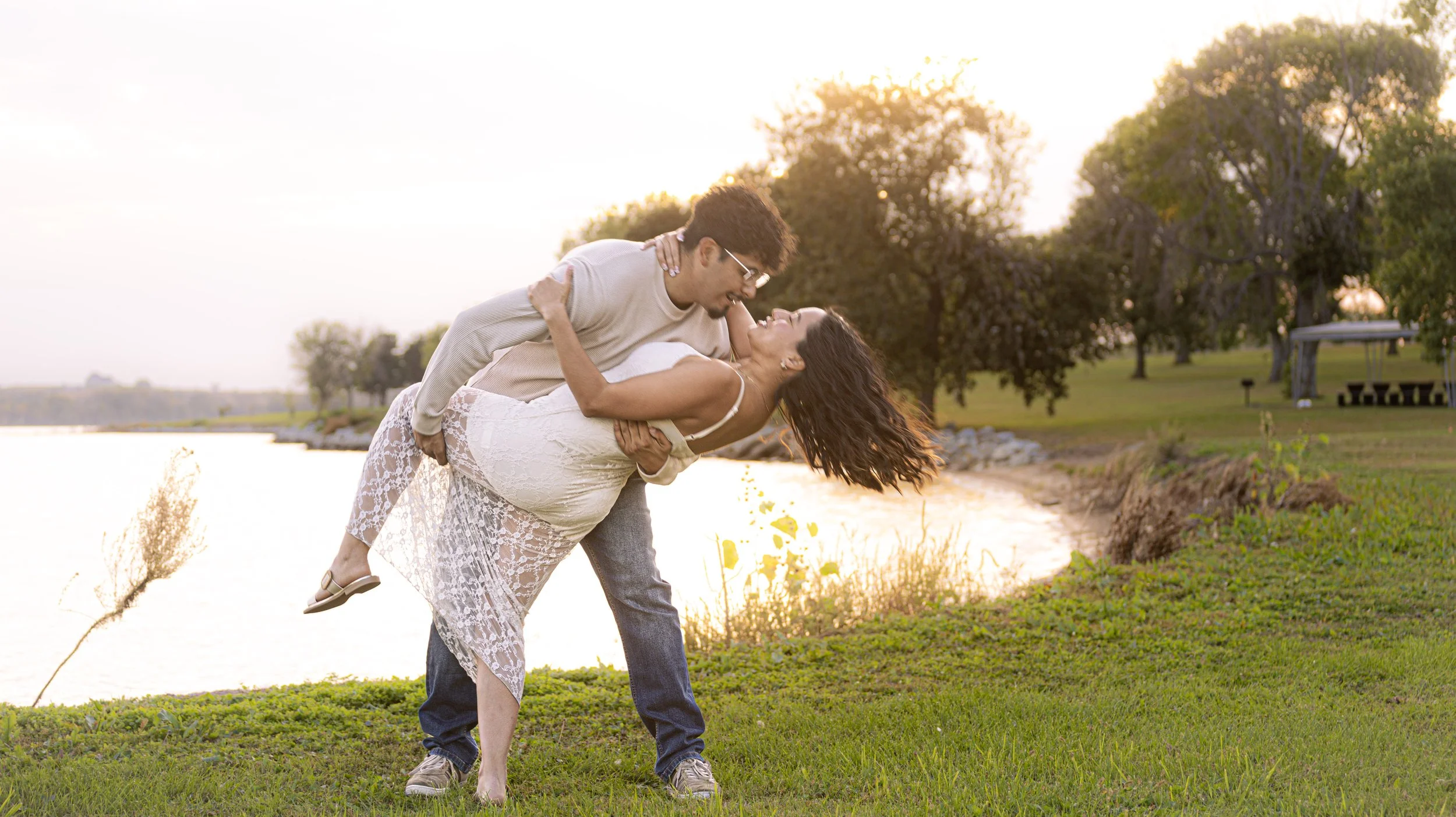 A man in glasses and a beige long-sleeve shirt dips a woman in a white lace dress by a lake at sunset, with trees and a grassy park in the background.