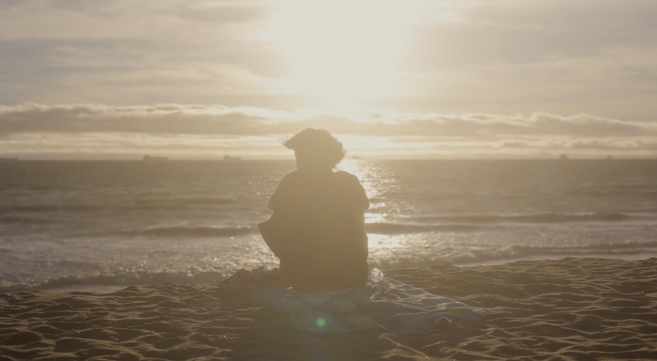 Person sitting on the beach near the ocean during sunset, facing the water with the sun low in the sky.