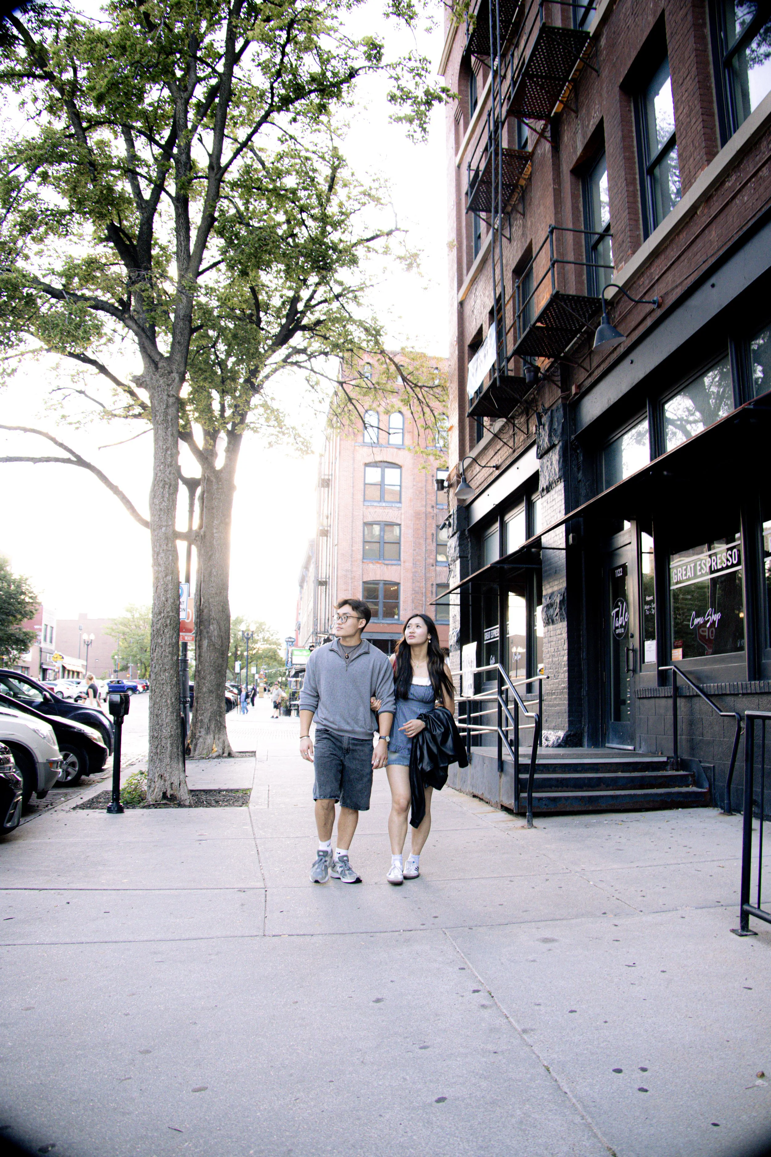 Two young people, a man and a woman, walking arm in arm on a city sidewalk with parked cars, trees, and brick buildings in the background during late afternoon.