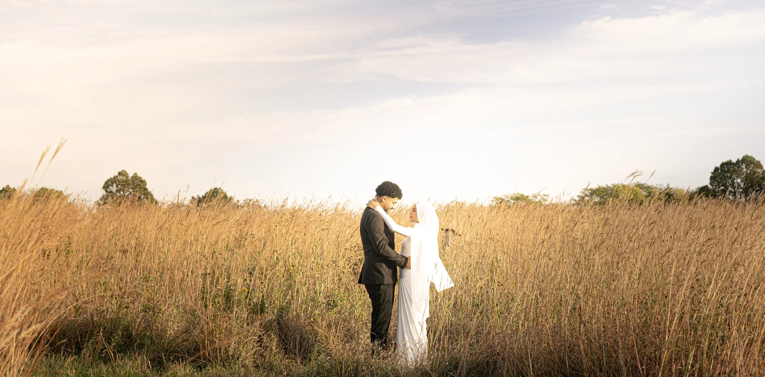 A couple standing in a field of tall grass during sunset, with the woman wearing a white dress and headscarf, and the man in a black suit, looking at each other lovingly.