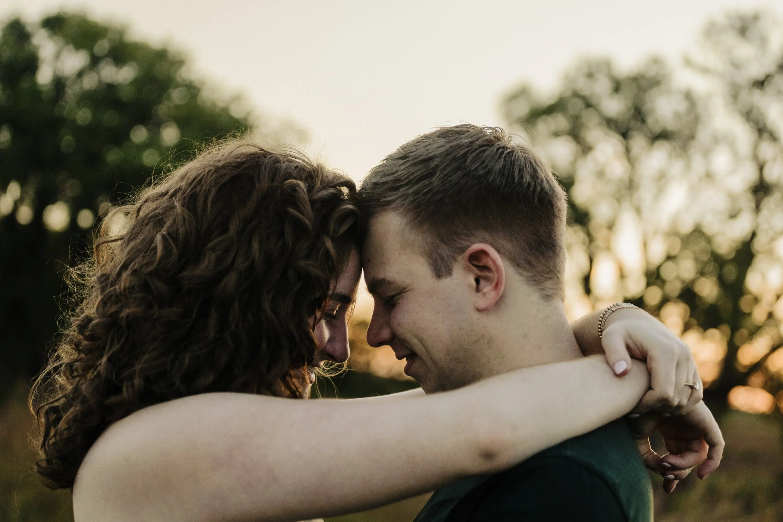 A couple embraces with foreheads touching during sunset, outdoors with trees in the background.