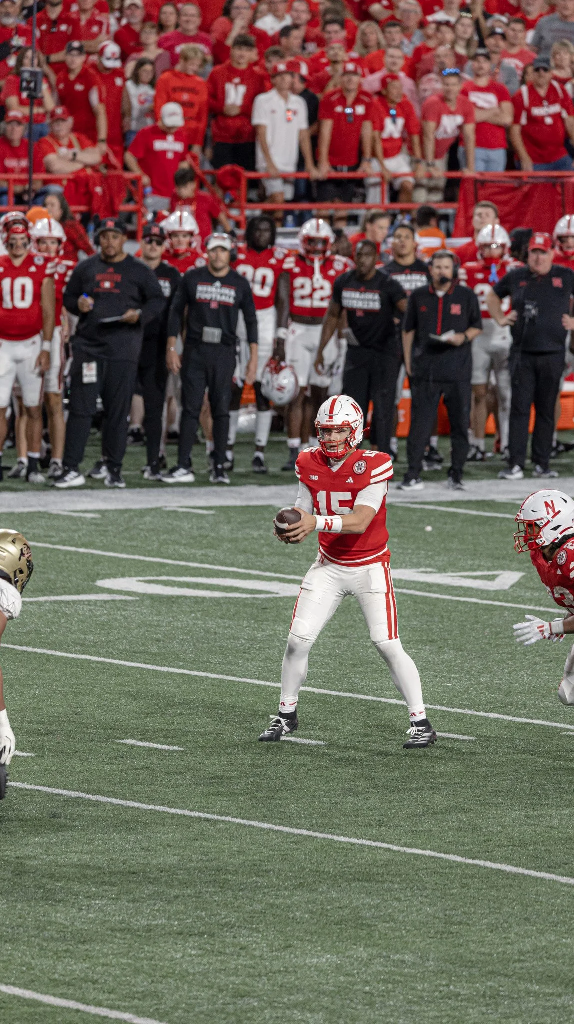 An American football game with a player wearing a red and white uniform with the number 15, holding the football and preparing to throw, while other players and coaches look on from the sideline, and a crowd of spectators in red shirts in the backgro
