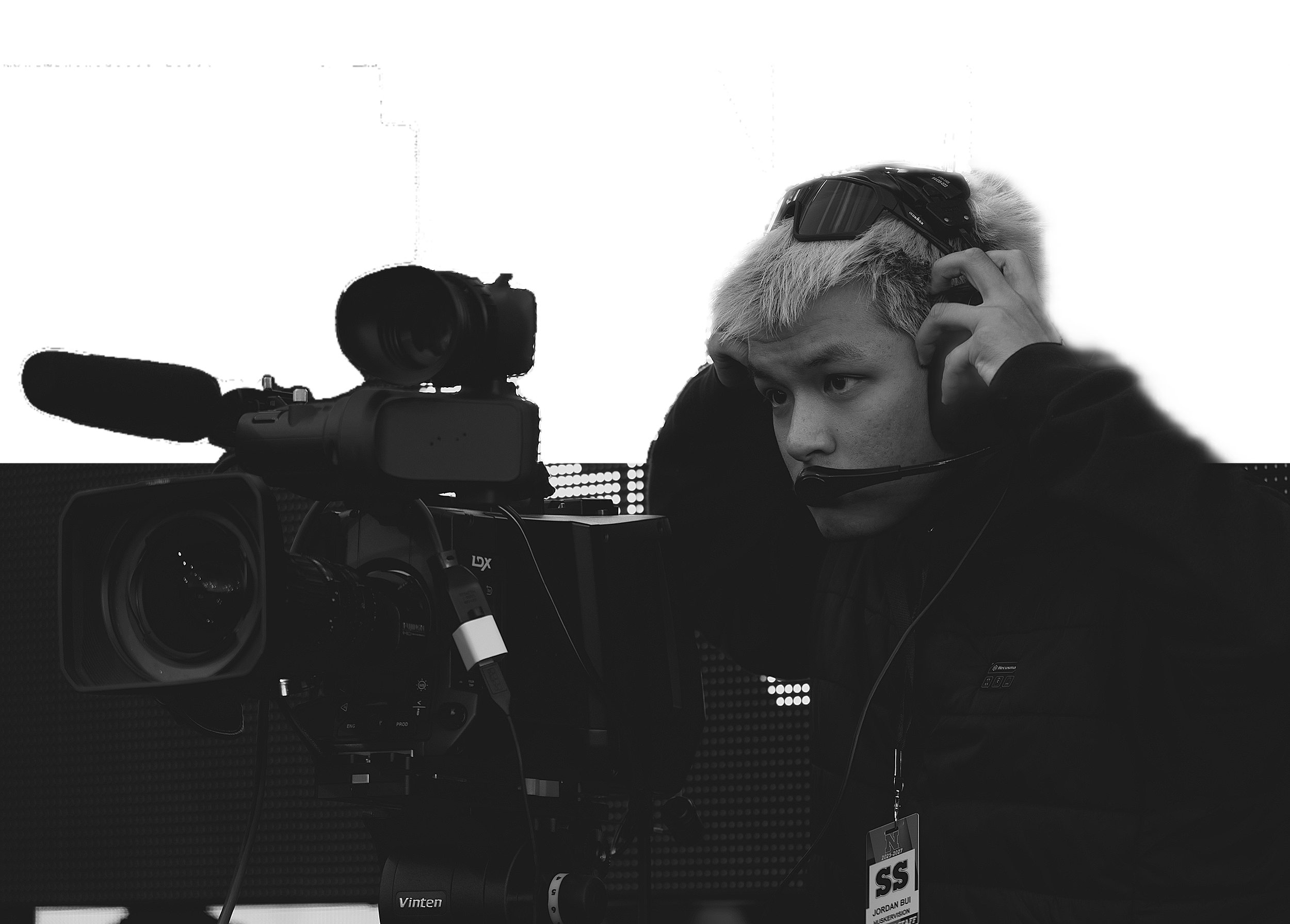 A young man with light-colored hair adjusts a headset while operating a professional video camera with a microphone attached, in a media setting.