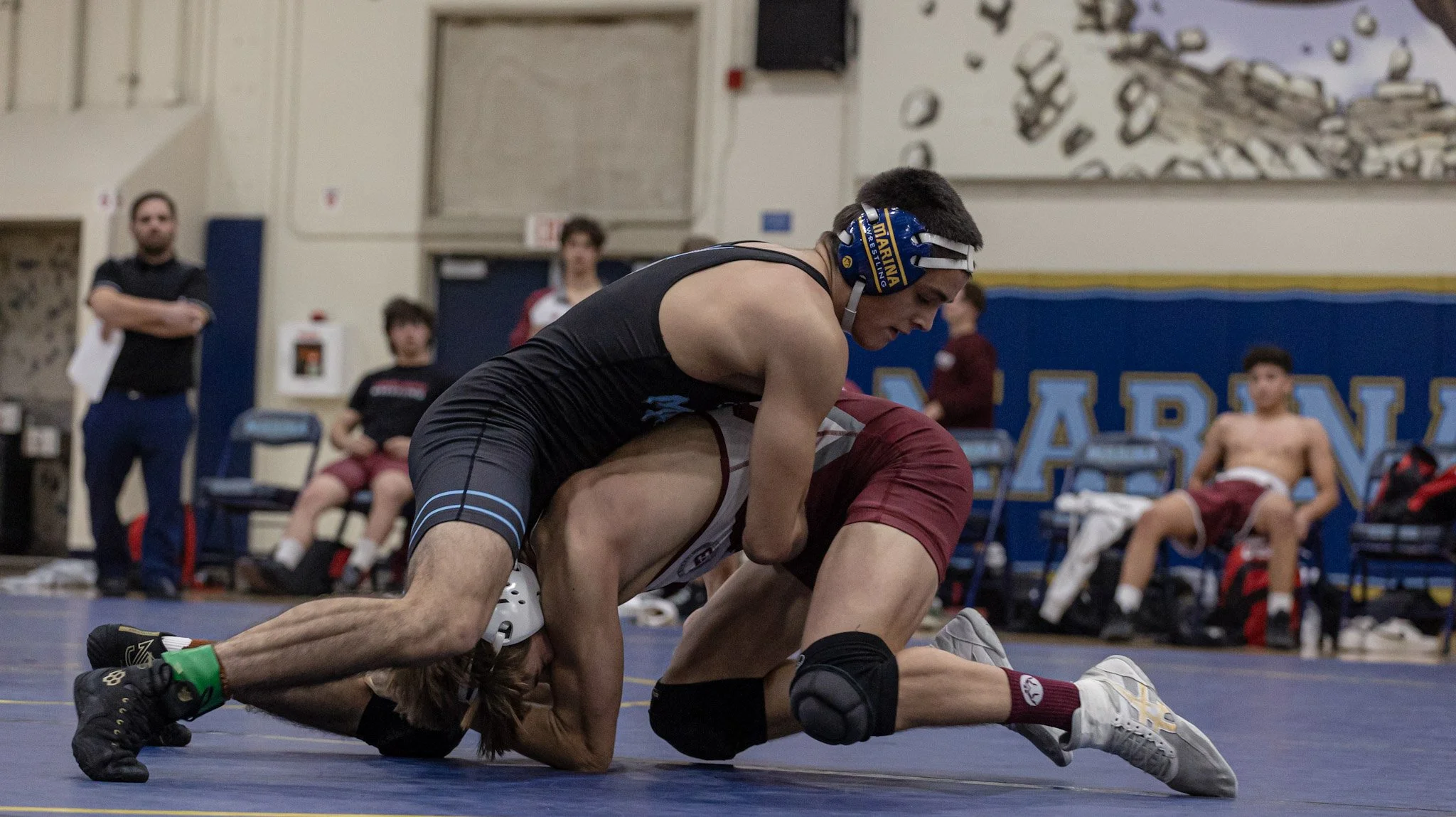 Two wrestlers compete on a blue wrestling mat, with one in a black singlet and the other in a maroon singlet, during a match. The wrestler in black is on top, trying to pin the opponent in maroon, who is on his hands and knees. In the background, sev