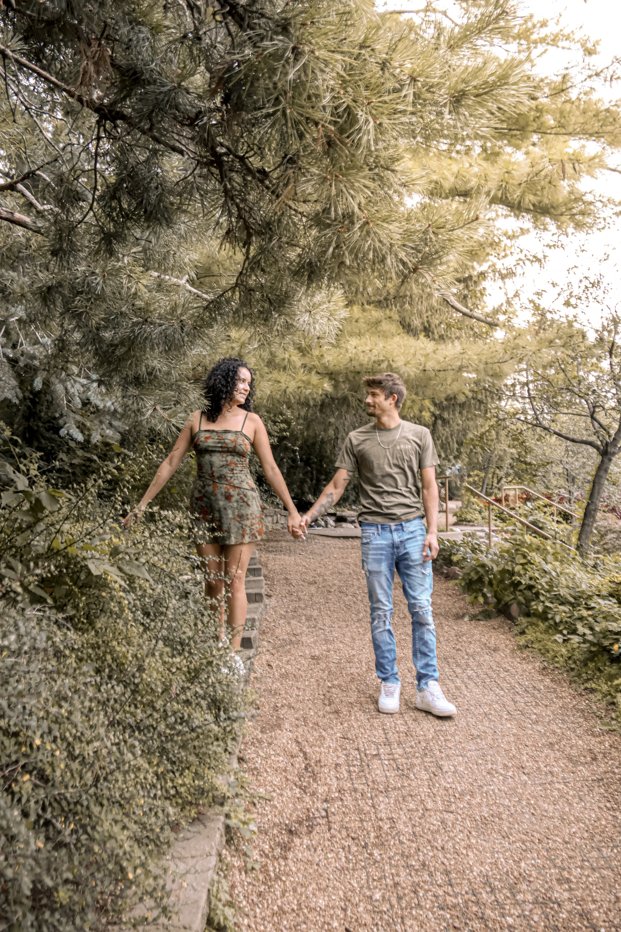 A young couple holding hands and walking along a garden path surrounded by trees and greenery.