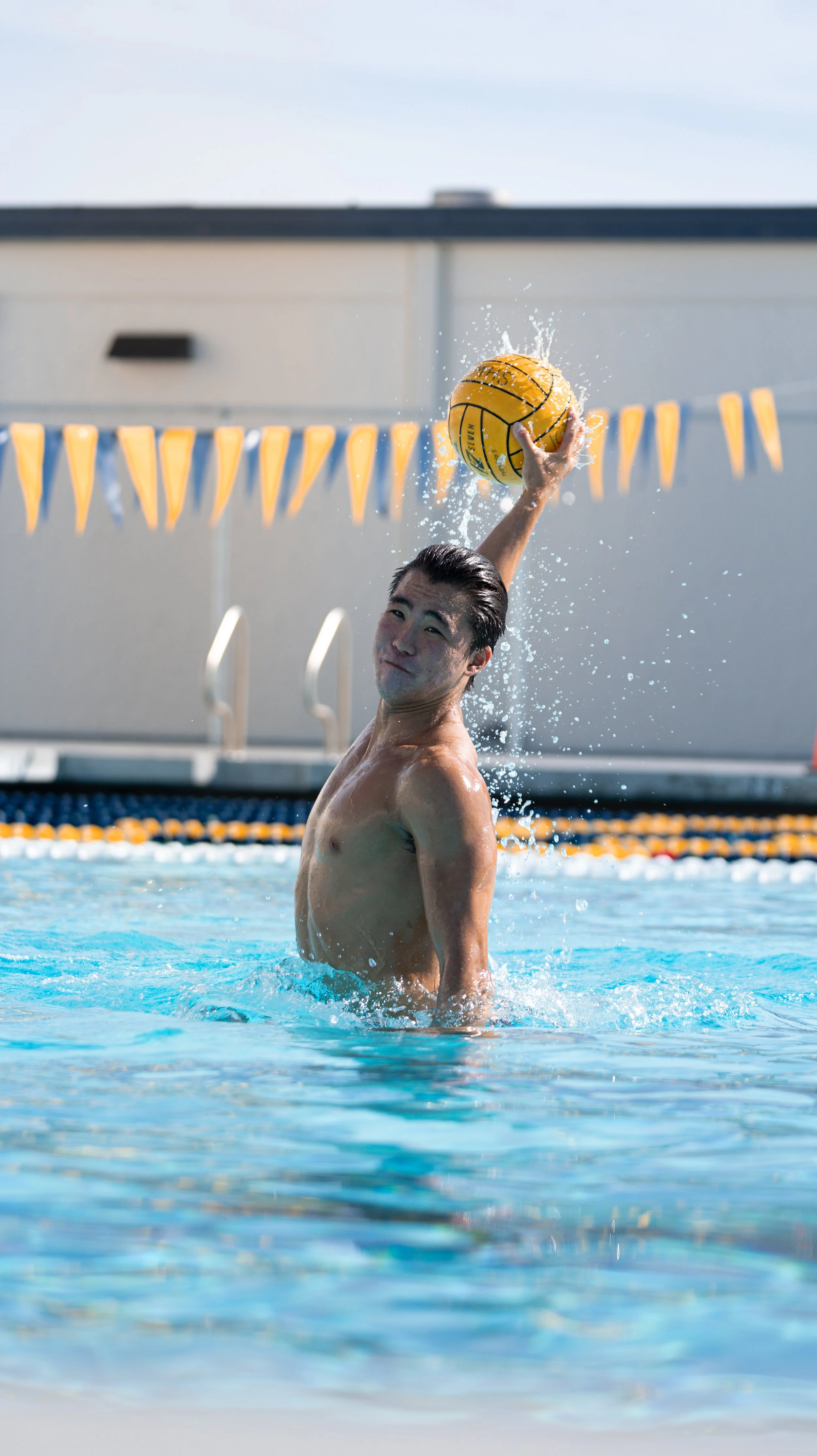 A shirtless man playing water polo in a swimming pool, holding a yellow water polo ball above his head.