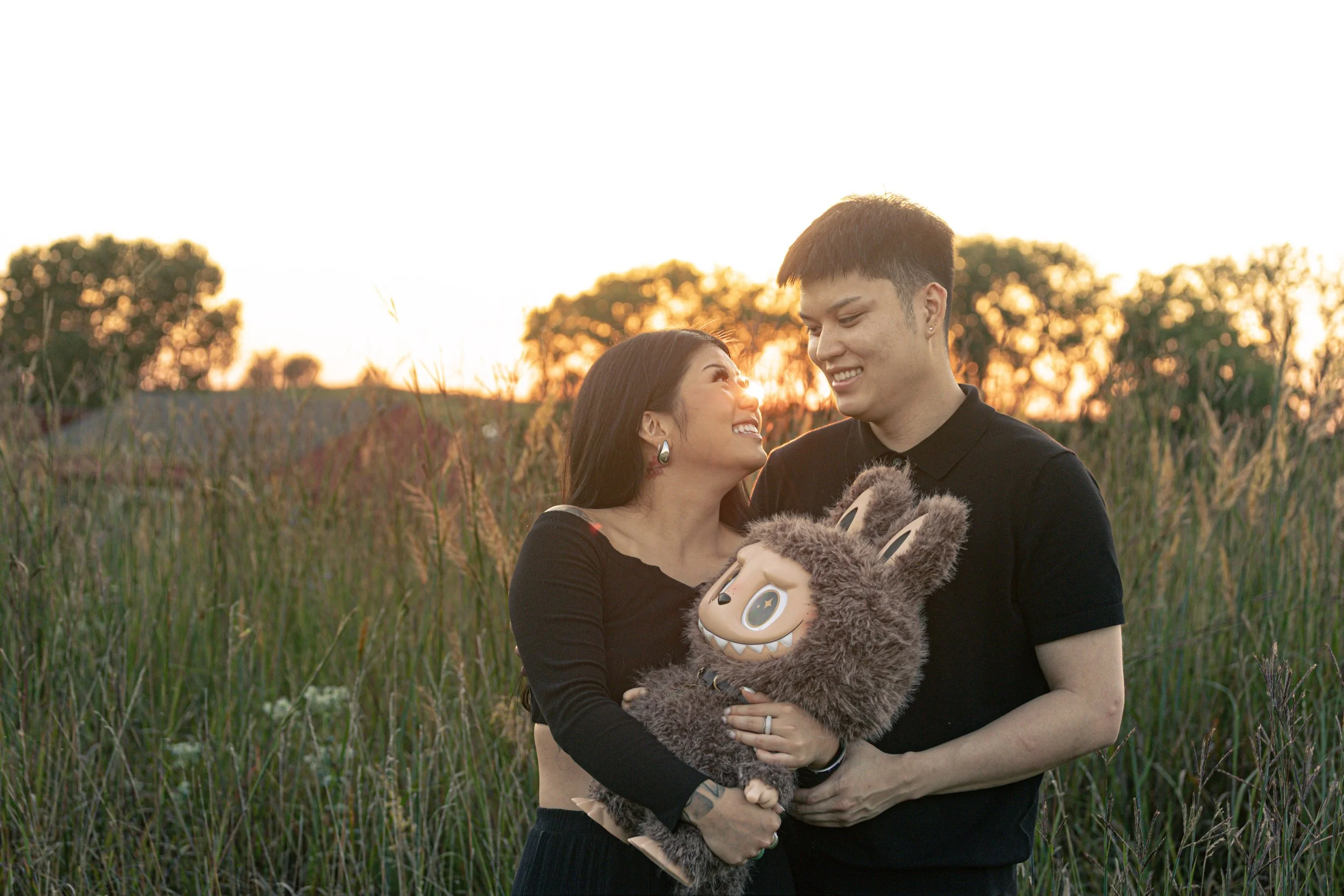 A woman and a man are standing in a grassy field during sunset, smiling at each other. The woman is holding a fluffy costume or puppet with a cartoonish face that has large eyes and sharp teeth.